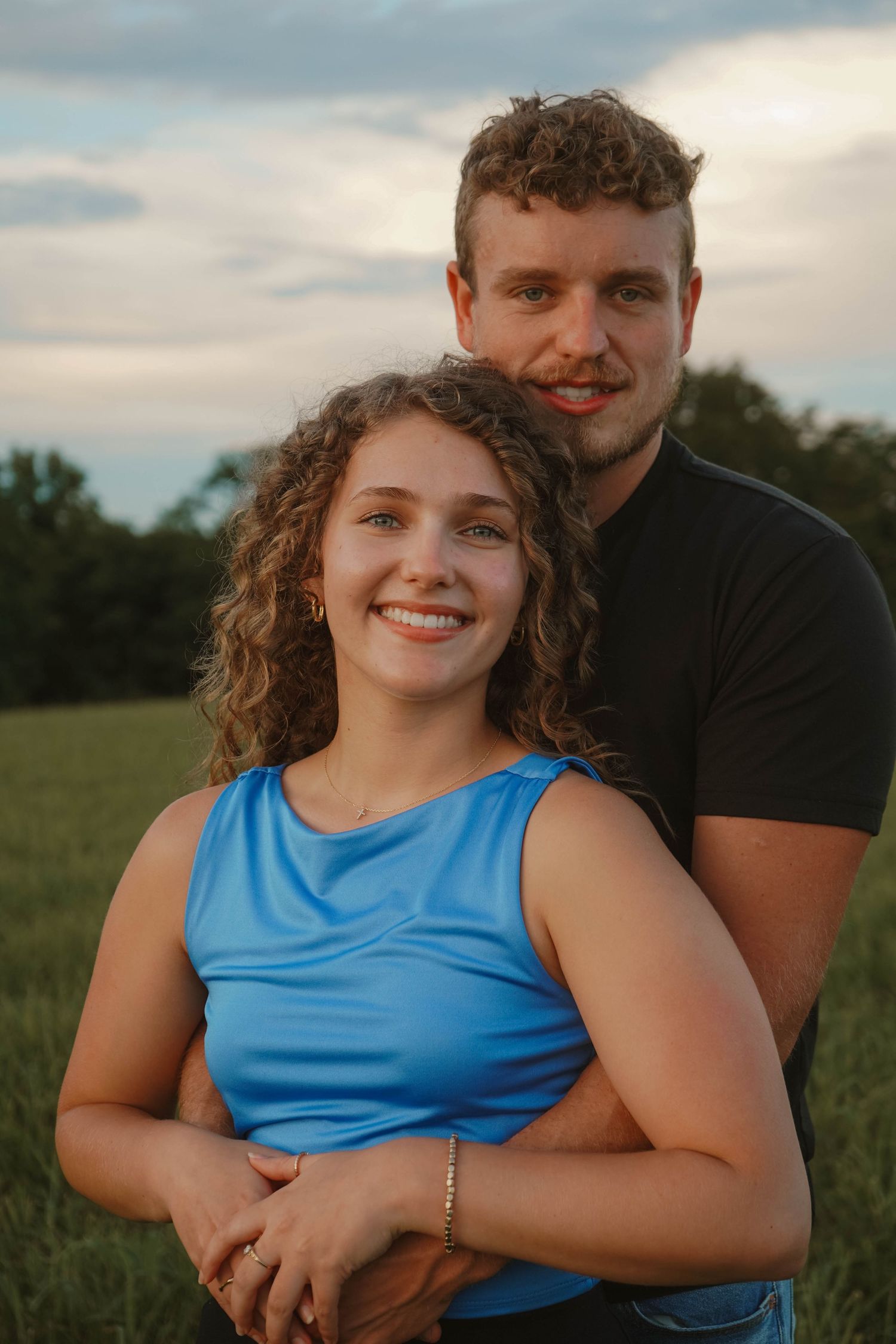 Smiling couple posing together in green field at dusk, man embracing woman from behind.