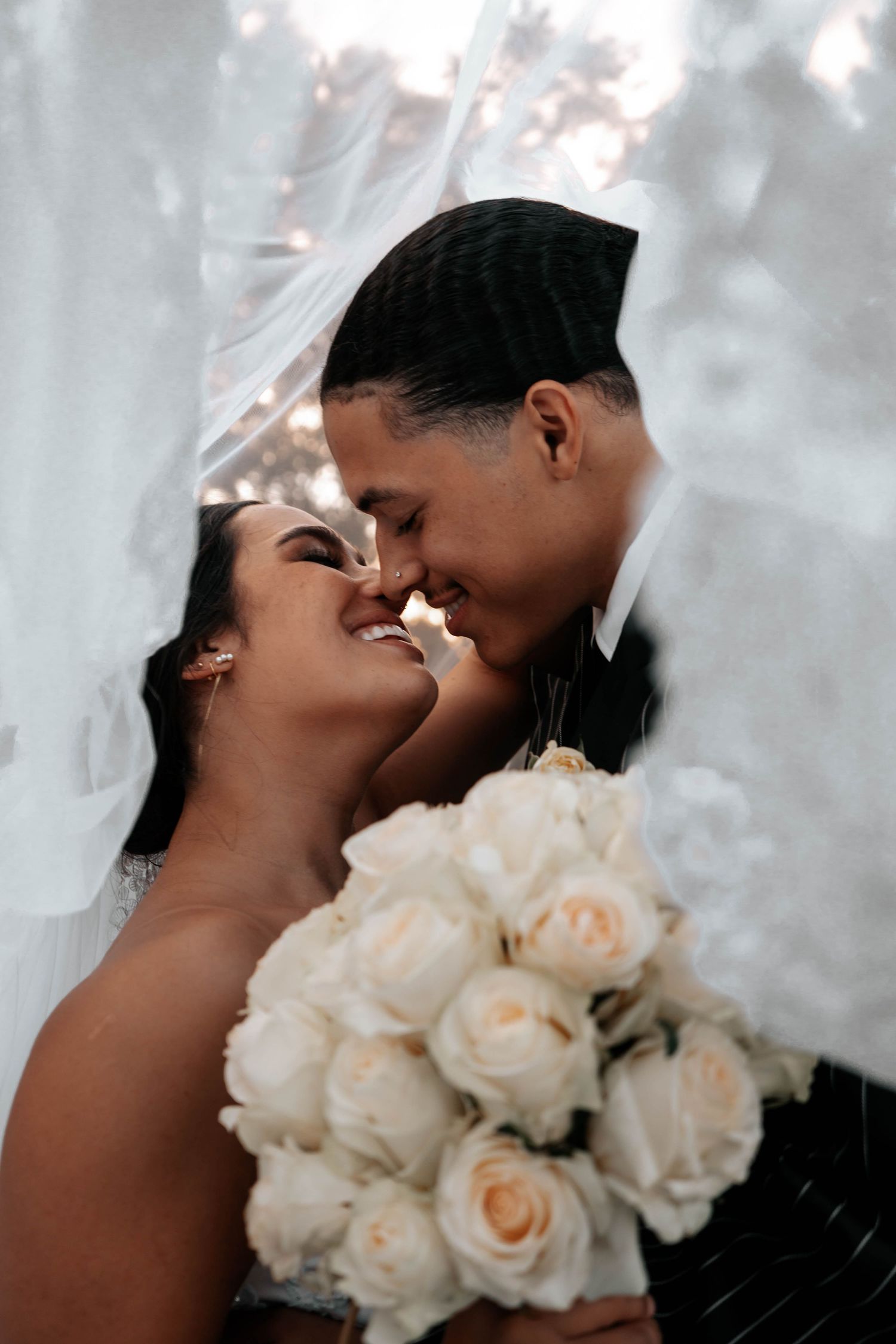 Bride and groom share intimate nose-to-nose moment under veil with white rose bouquet at wedding.