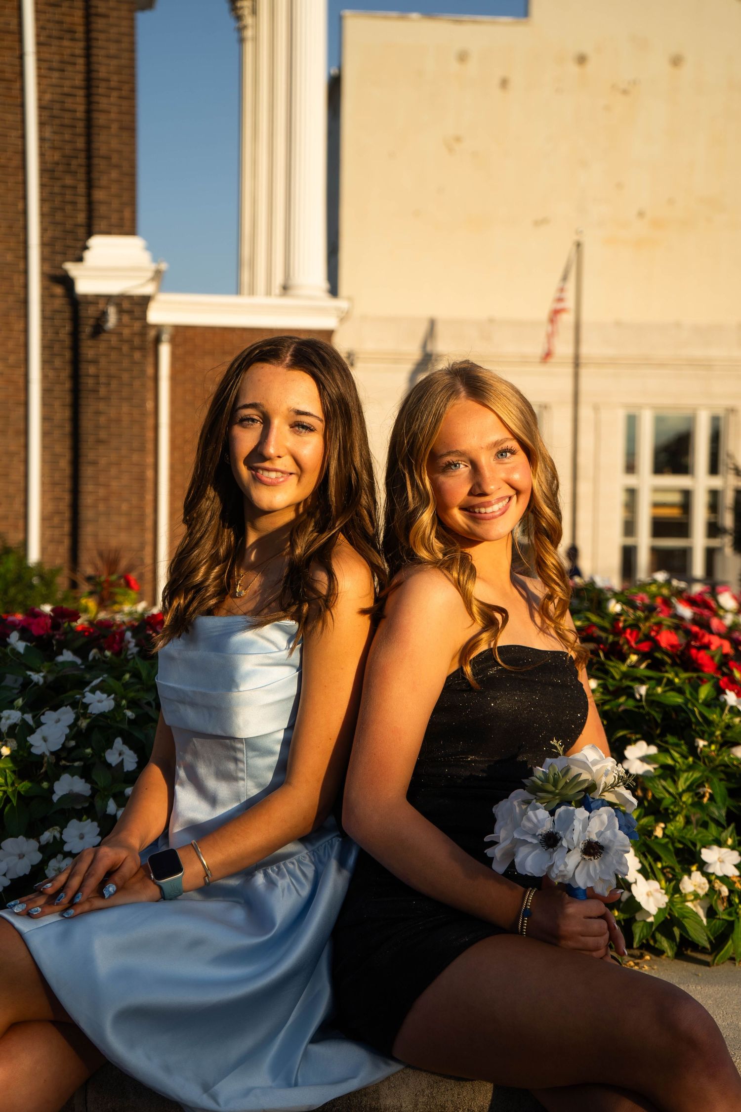 Two teenage girls in formal dresses sit back-to-back among colorful flowers outside brick building.