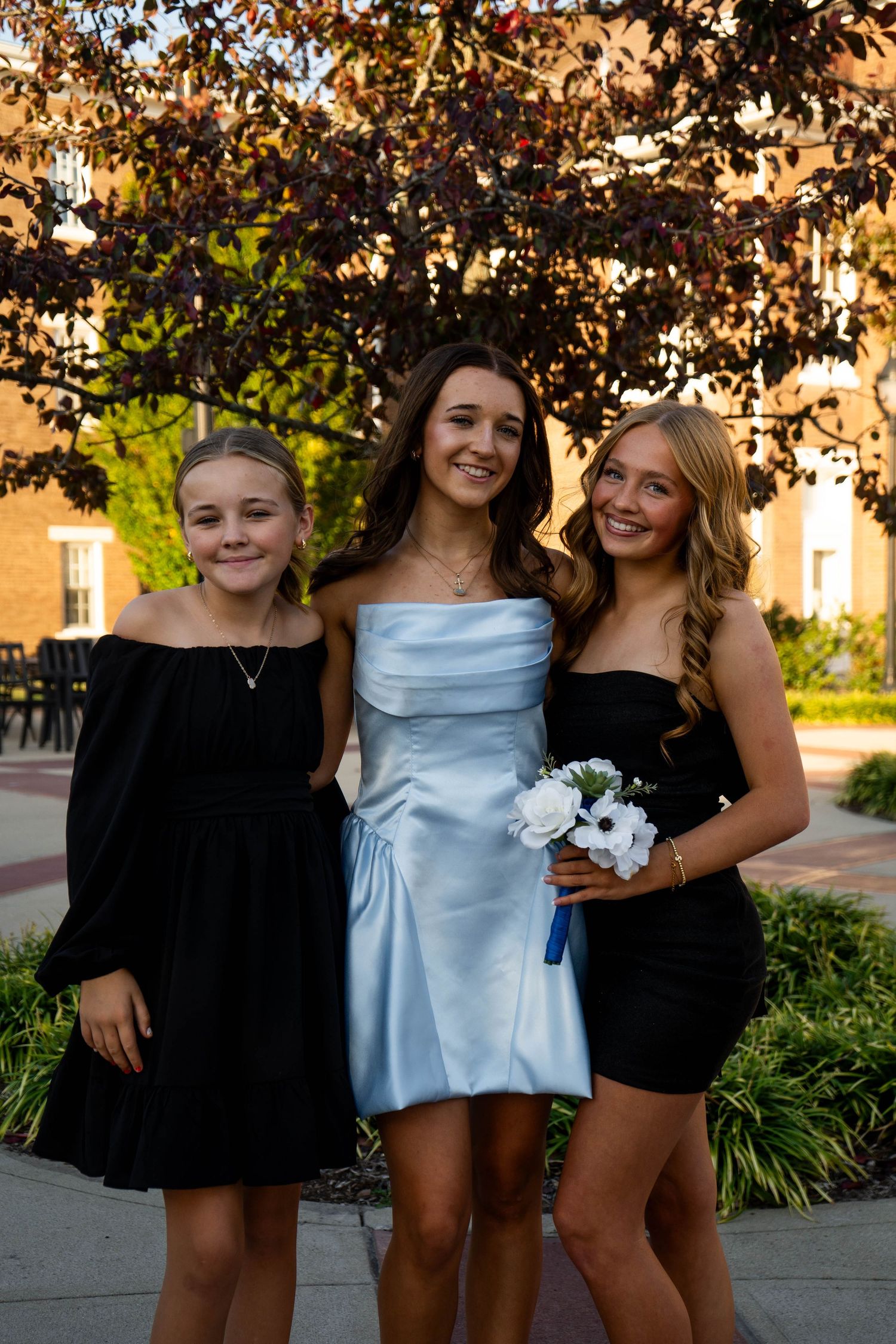 Three girls pose together under a purple-leafed tree, with the middle girl in a light blue strapless prom dress.