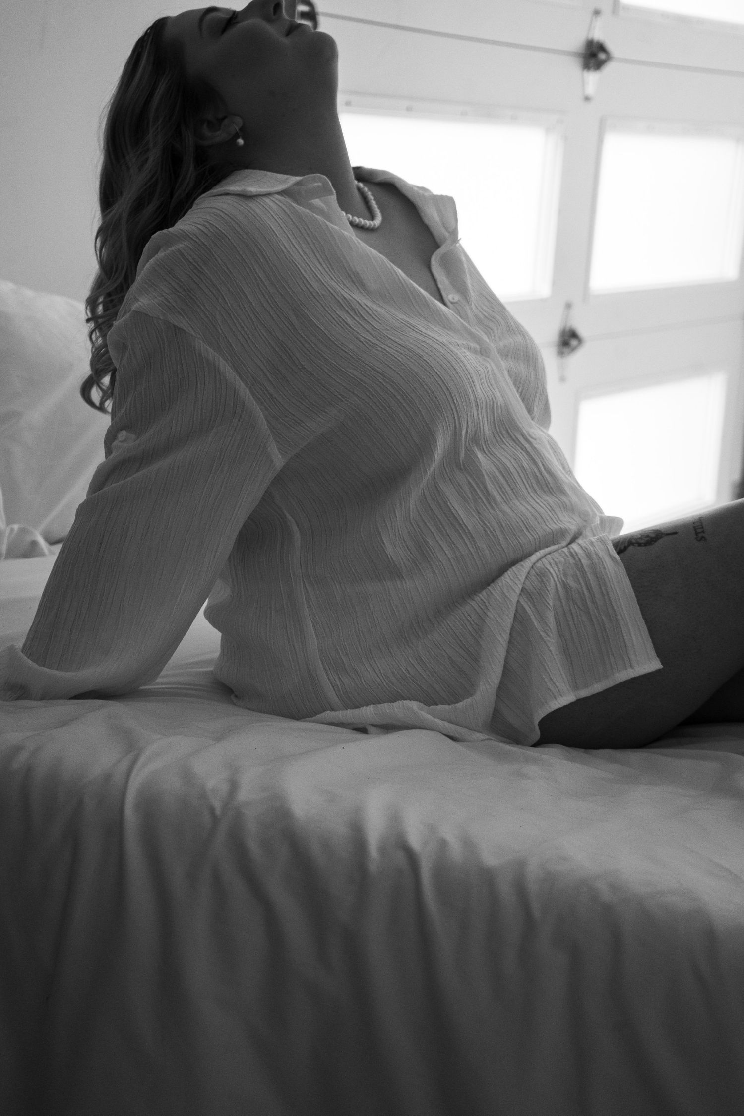 Black and white photo of a woman in a white linen shirt sitting on a bed near a bright window.
