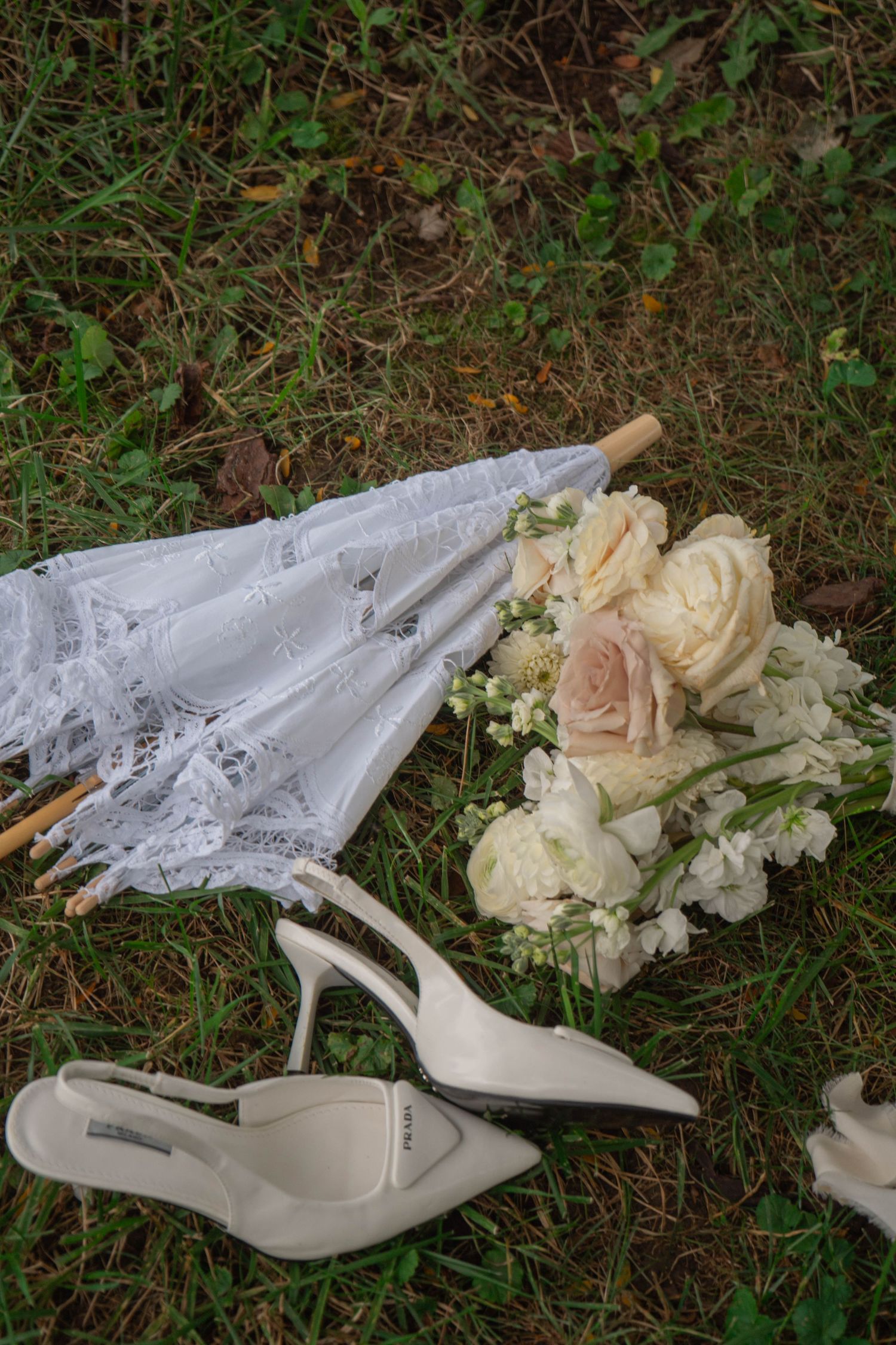 Bridal accessories flat lay featuring white lace umbrella, bouquet of roses, and elegant heeled shoes on grass.