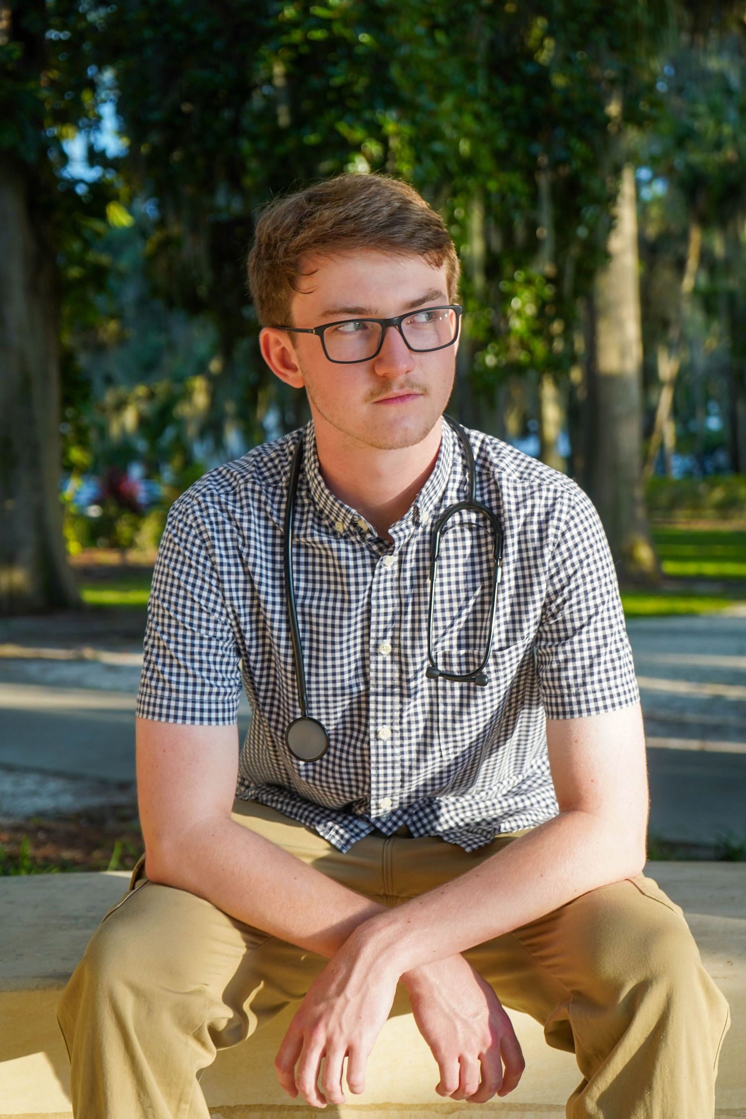 Thoughtful young man wearing glasses and stethoscope sitting outdoors in a tree-lined park setting.