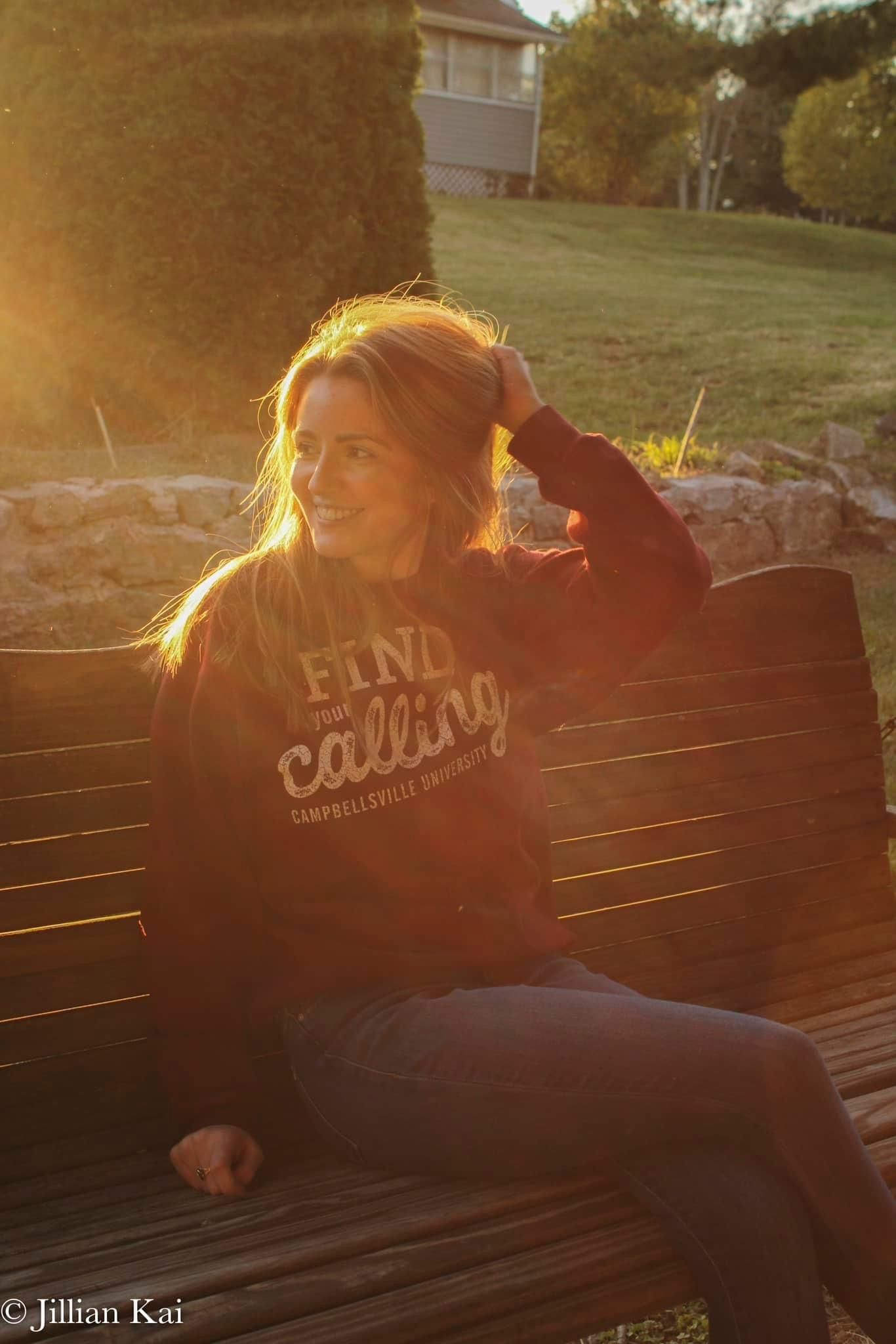 Blonde woman in Campbellsville University sweatshirt smiling on bench in warm golden hour sunlight.