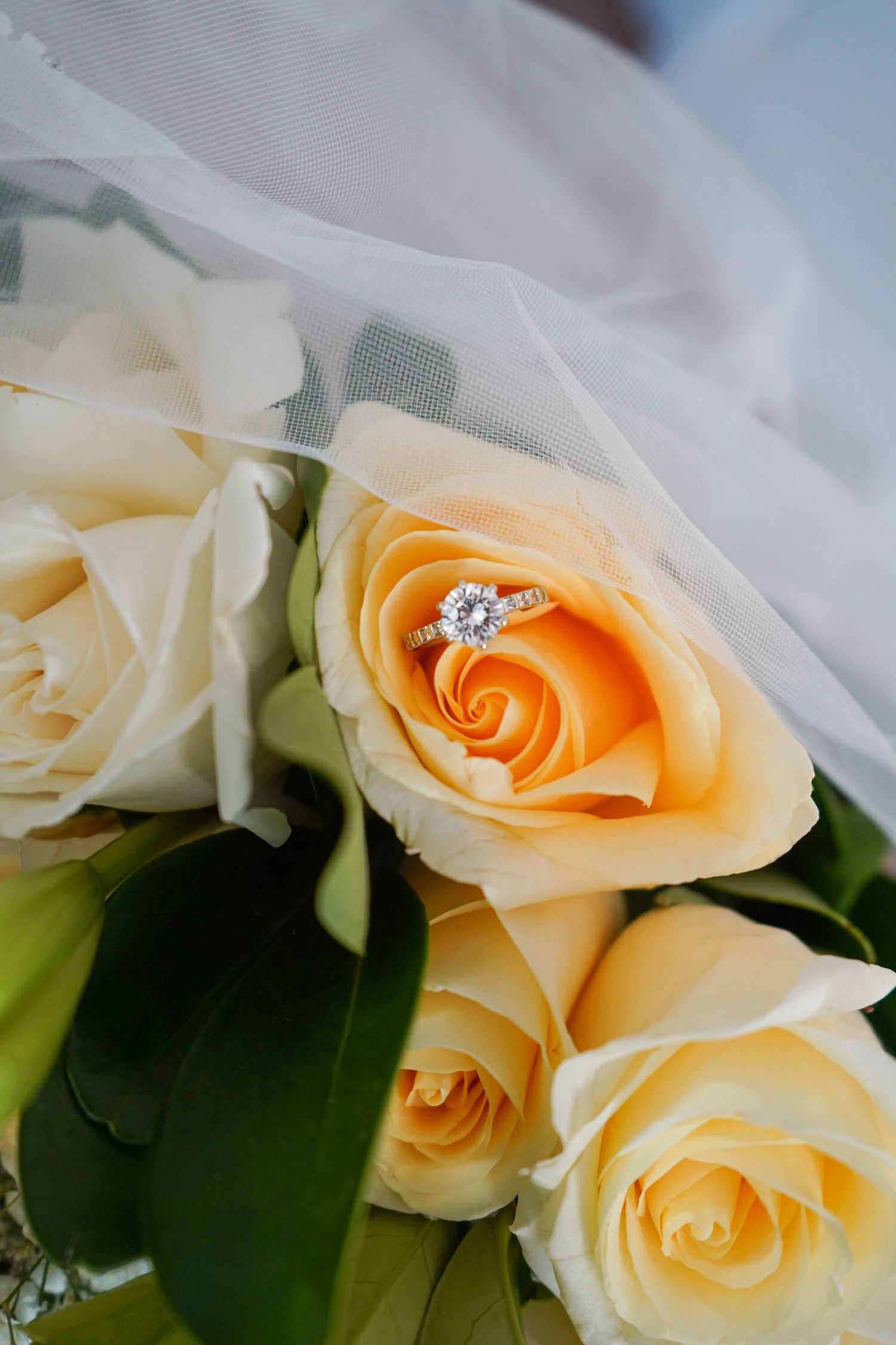 Diamond solitaire engagement ring resting inside a cream rose on a bridal bouquet with white tulle veil.