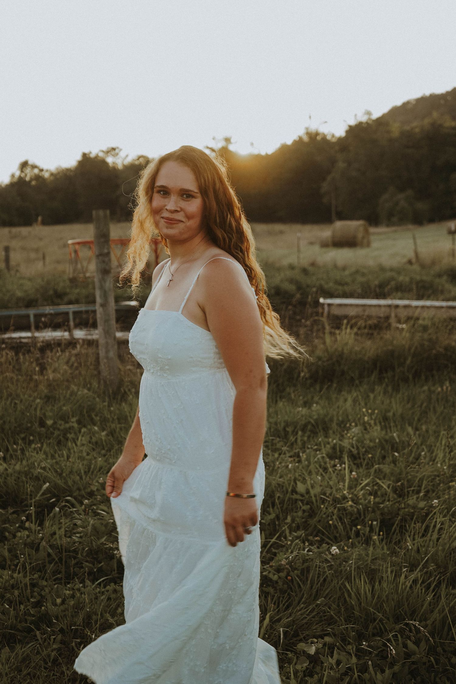 Smiling young woman in white dress posing in a sunlit farm field at golden hour.