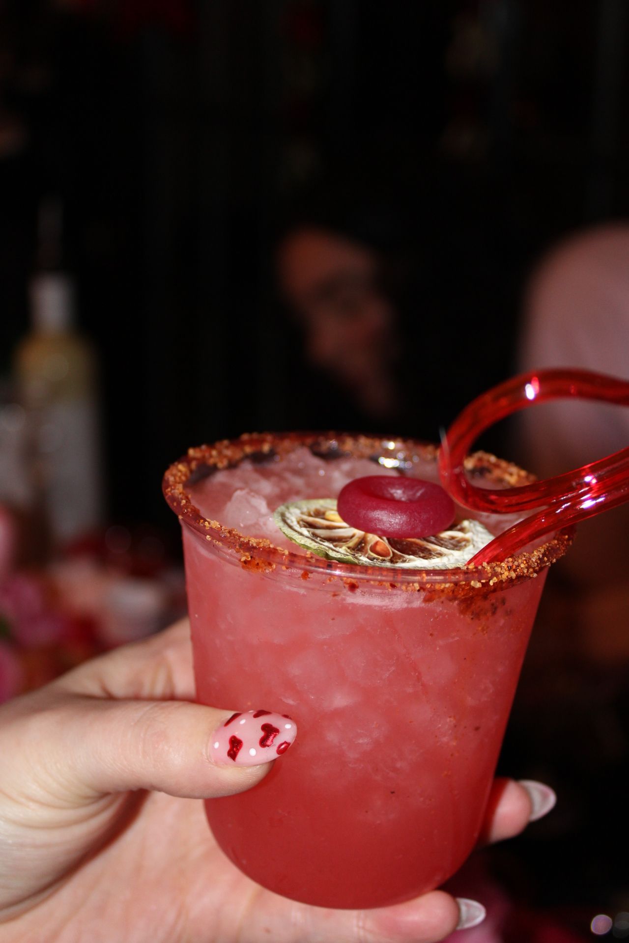 A hand with red nails holds a pink cocktail with a salted rim, red straw, and garnishes in a dimly lit bar.
