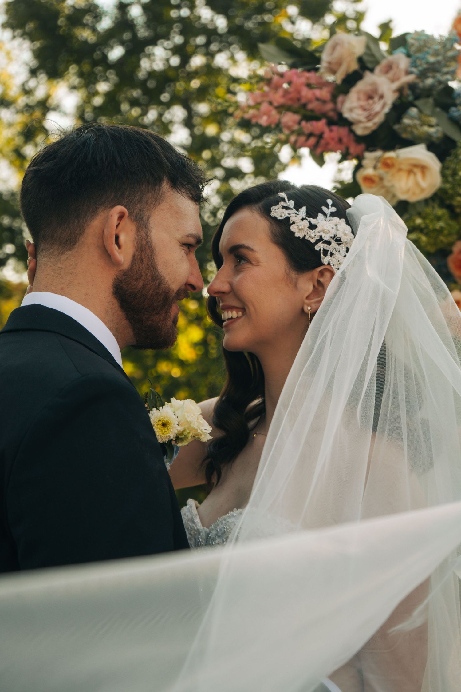 Smiling bride in veil and groom sharing intimate moment under floral arch.