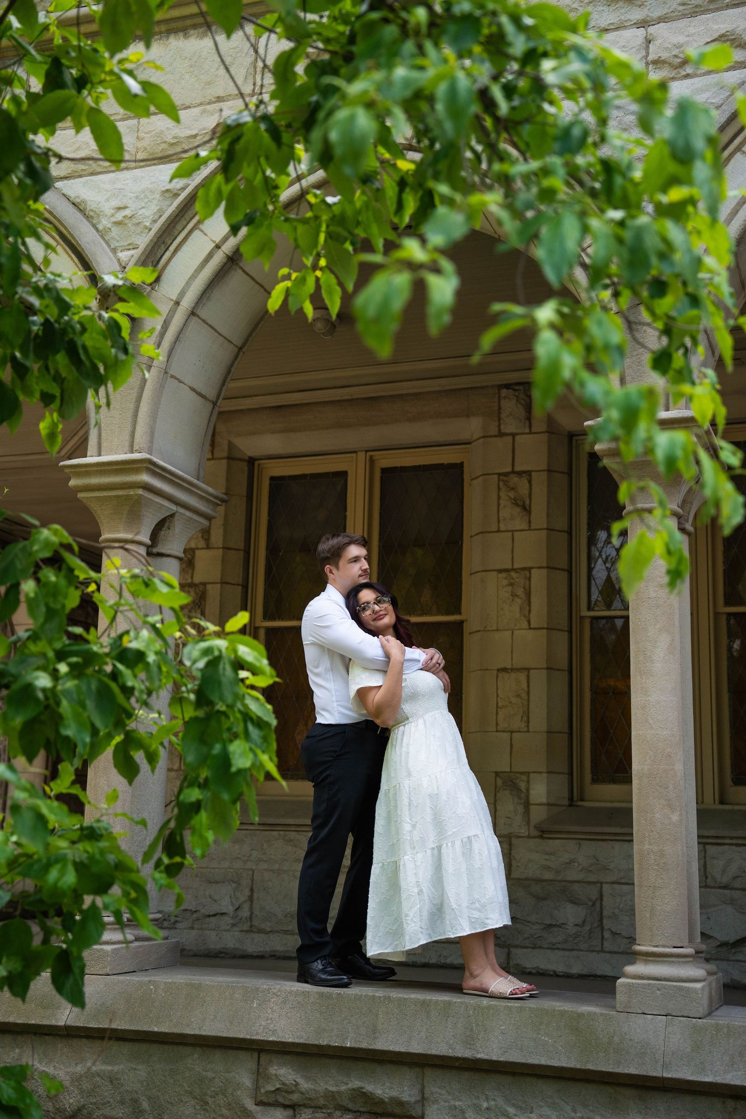 Couple embracing on stone church steps, man in white shirt and woman in white dress, framed by green leaves.