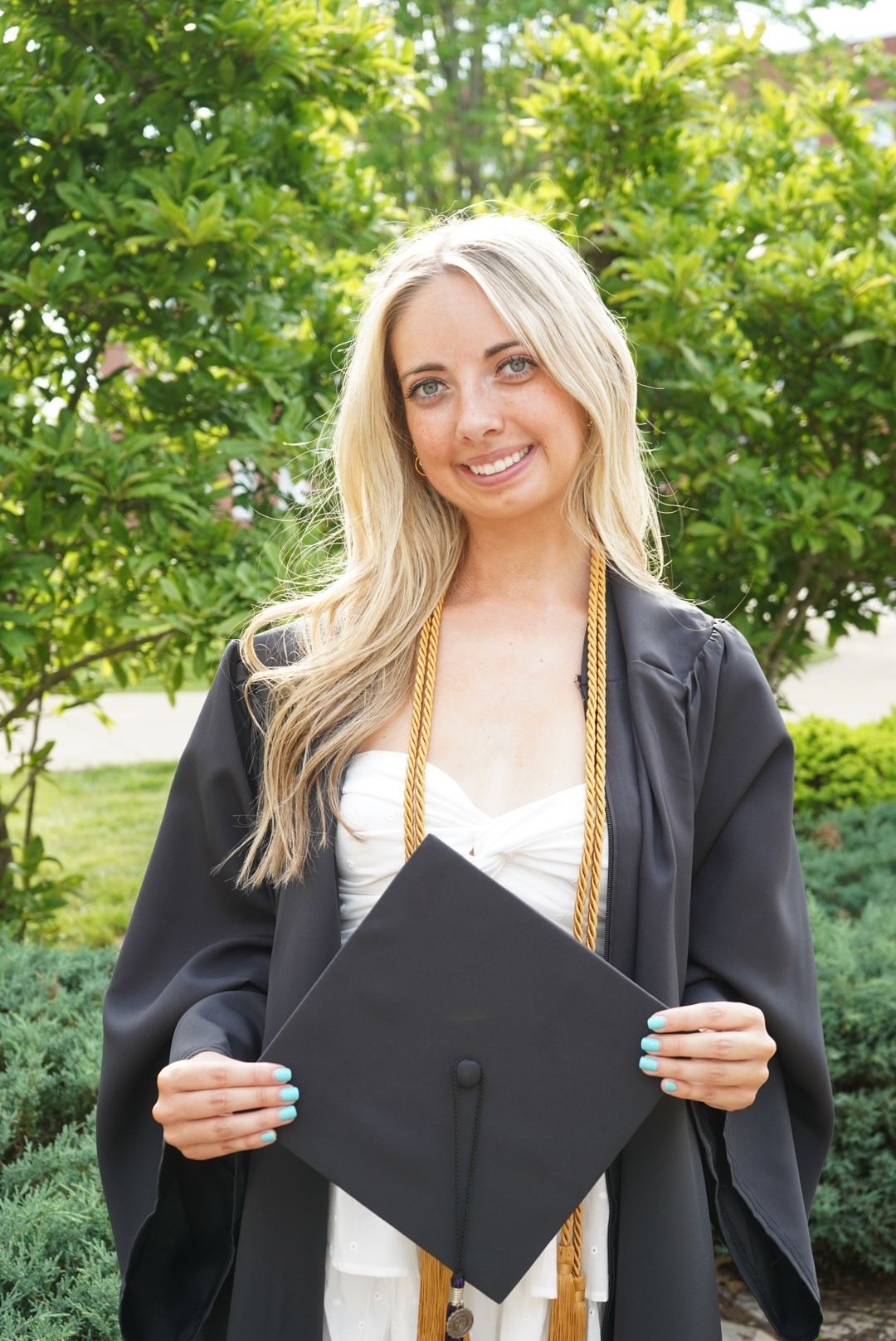 Blonde graduate smiling in black cap and gown with gold honor cord outdoors.