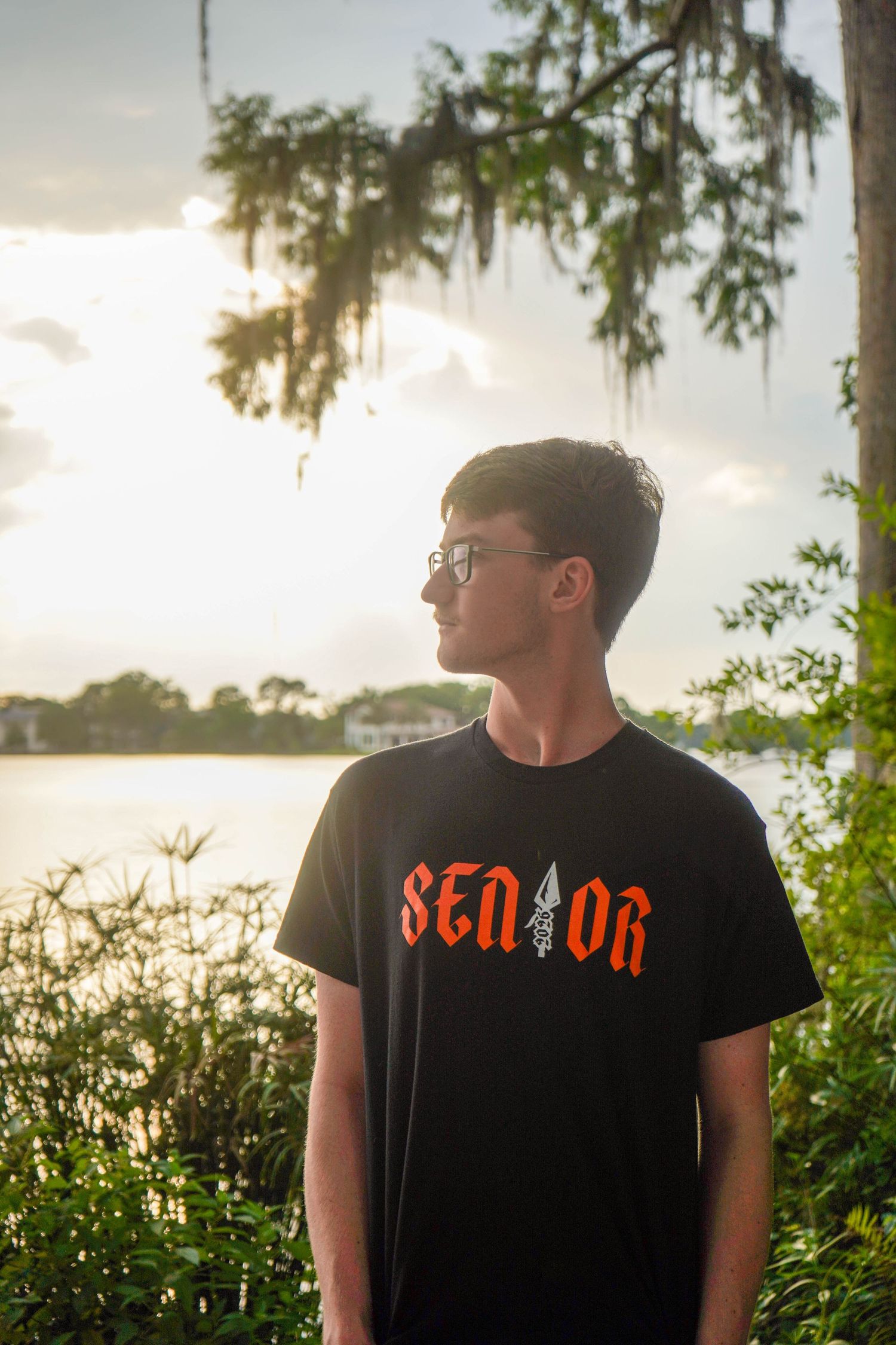 Young man in black Senior t-shirt standing near lake with Spanish moss trees.