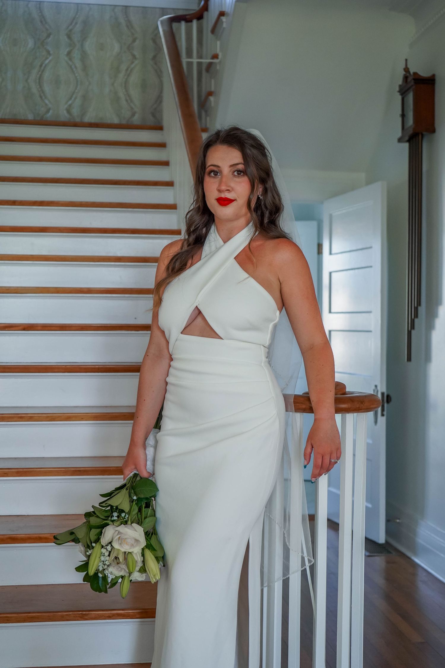 Beautiful bride in white crossover halter wedding gown with cutout detail poses on wooden staircase holding bouquet.