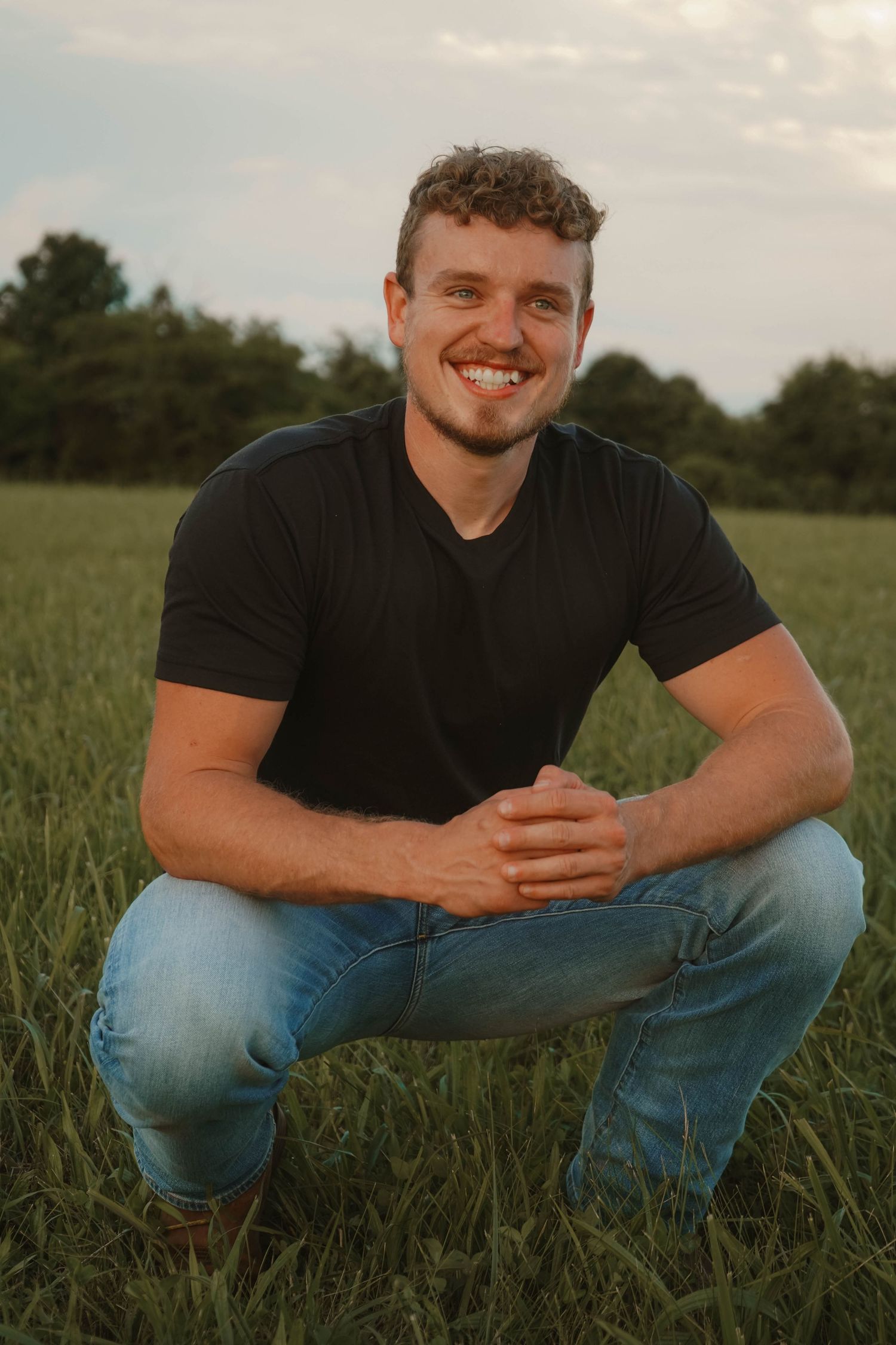 Smiling young man in black t-shirt crouches in lush green field at sunset.