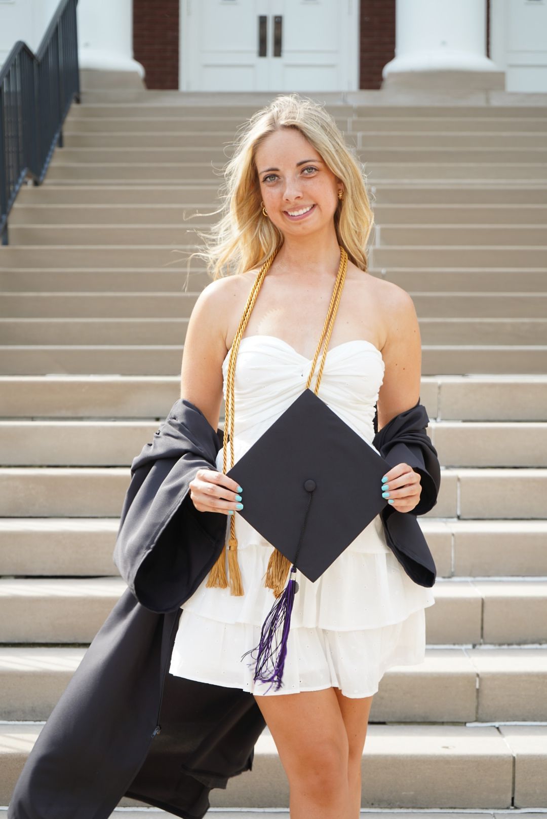 Smiling blonde graduate in cap and gown holding mortarboard on university steps.