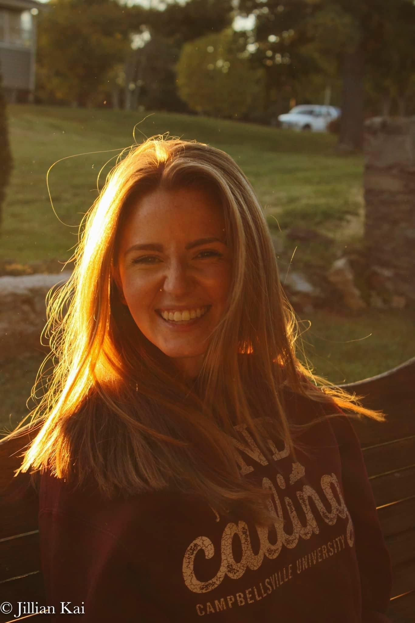 Smiling blonde woman in Campbellsville University sweatshirt bathed in golden sunset light outdoors.