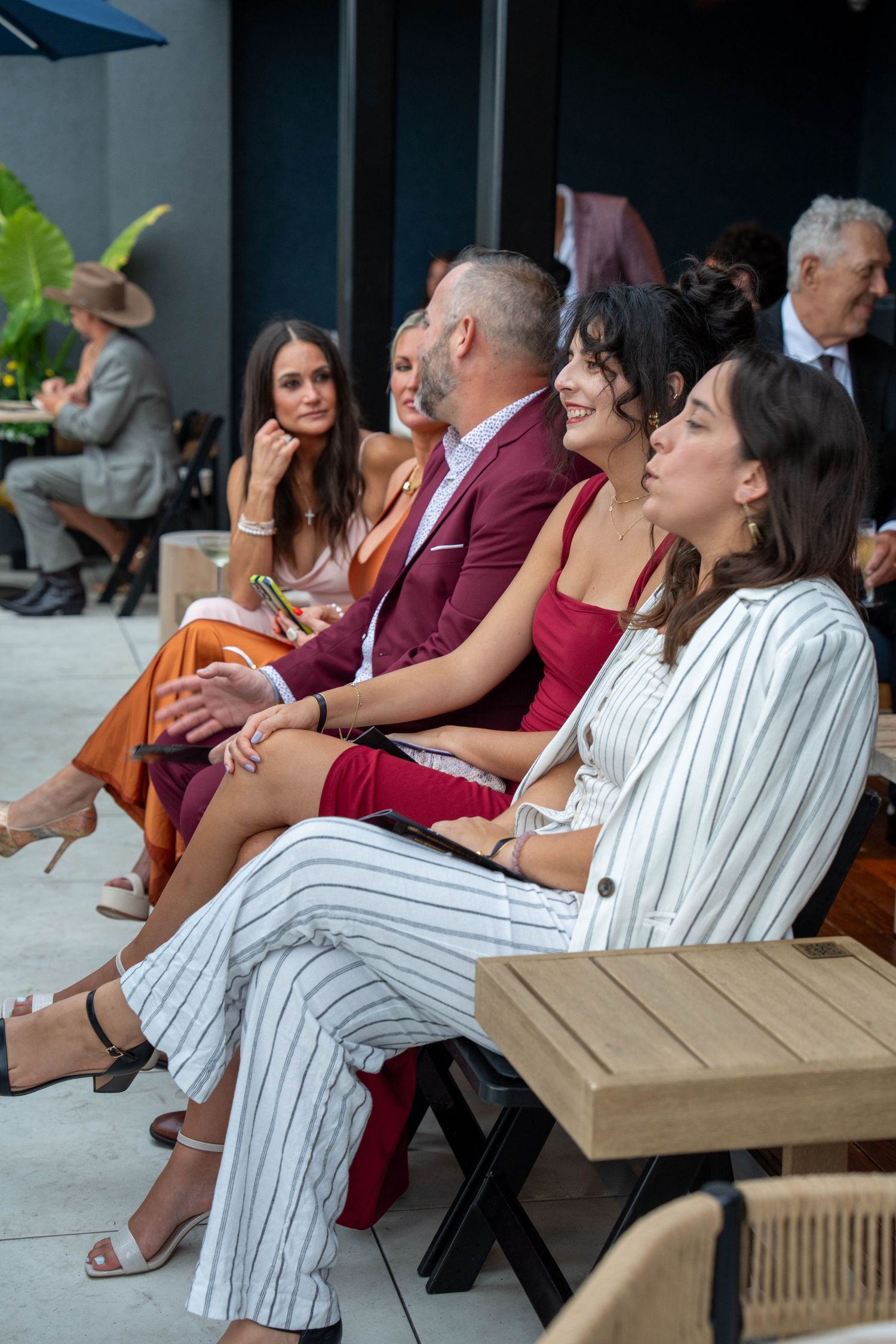 Elegantly dressed guests seated outdoors at a formal event, wearing colorful attire including red, orange, and striped outfits.