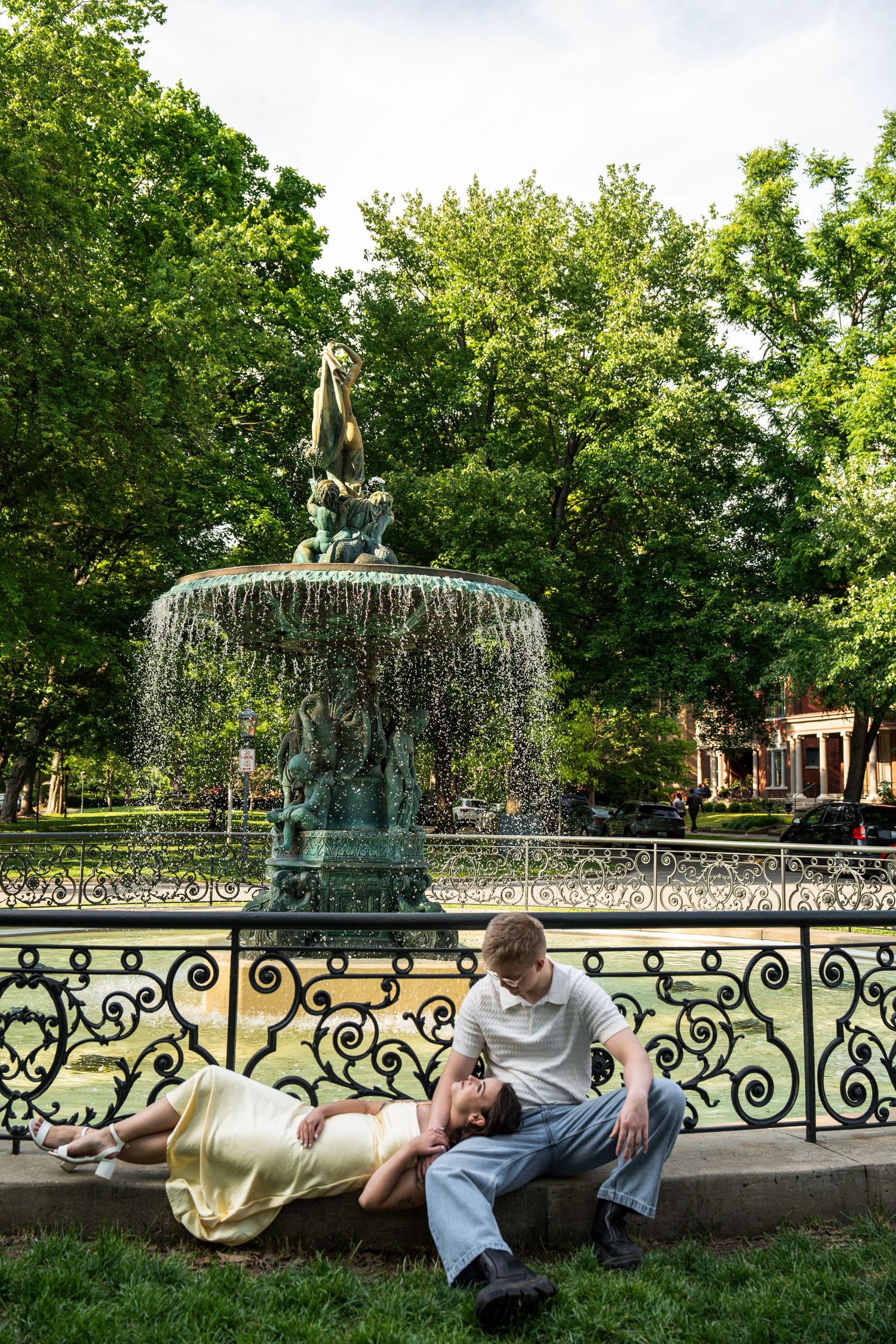 Couple relaxing by an ornate fountain in a green park, woman lying on grass.