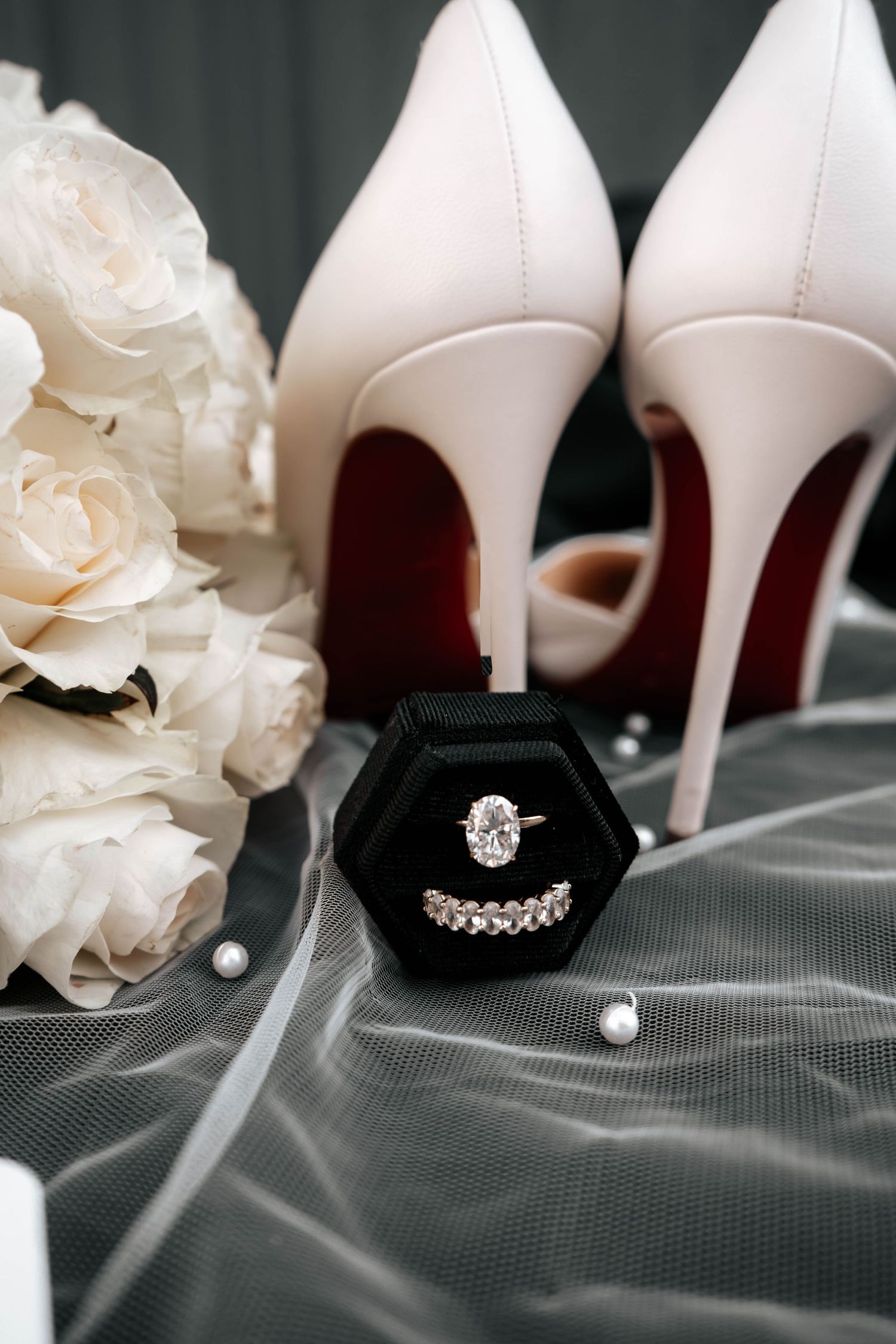 Bridal flat lay with diamond engagement ring and wedding band in black velvet box, white heels, and pearl accents.