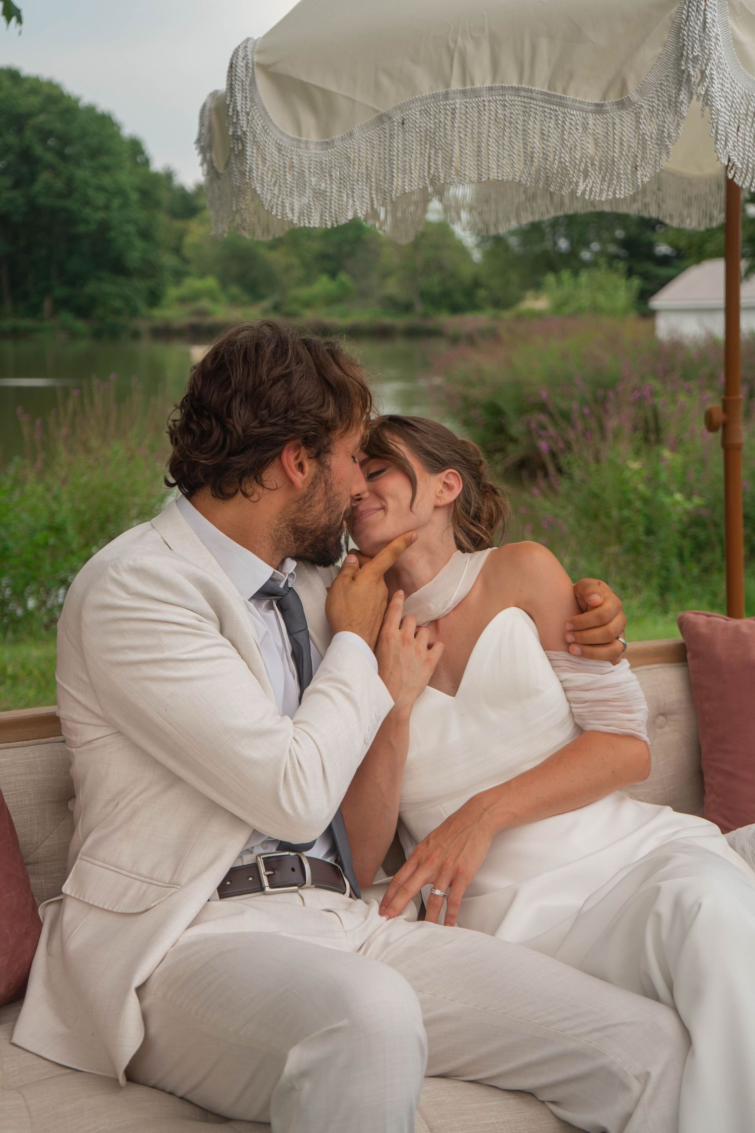Groom in white suit tenderly kisses bride on an outdoor sofa beneath a fringed umbrella near a pond.