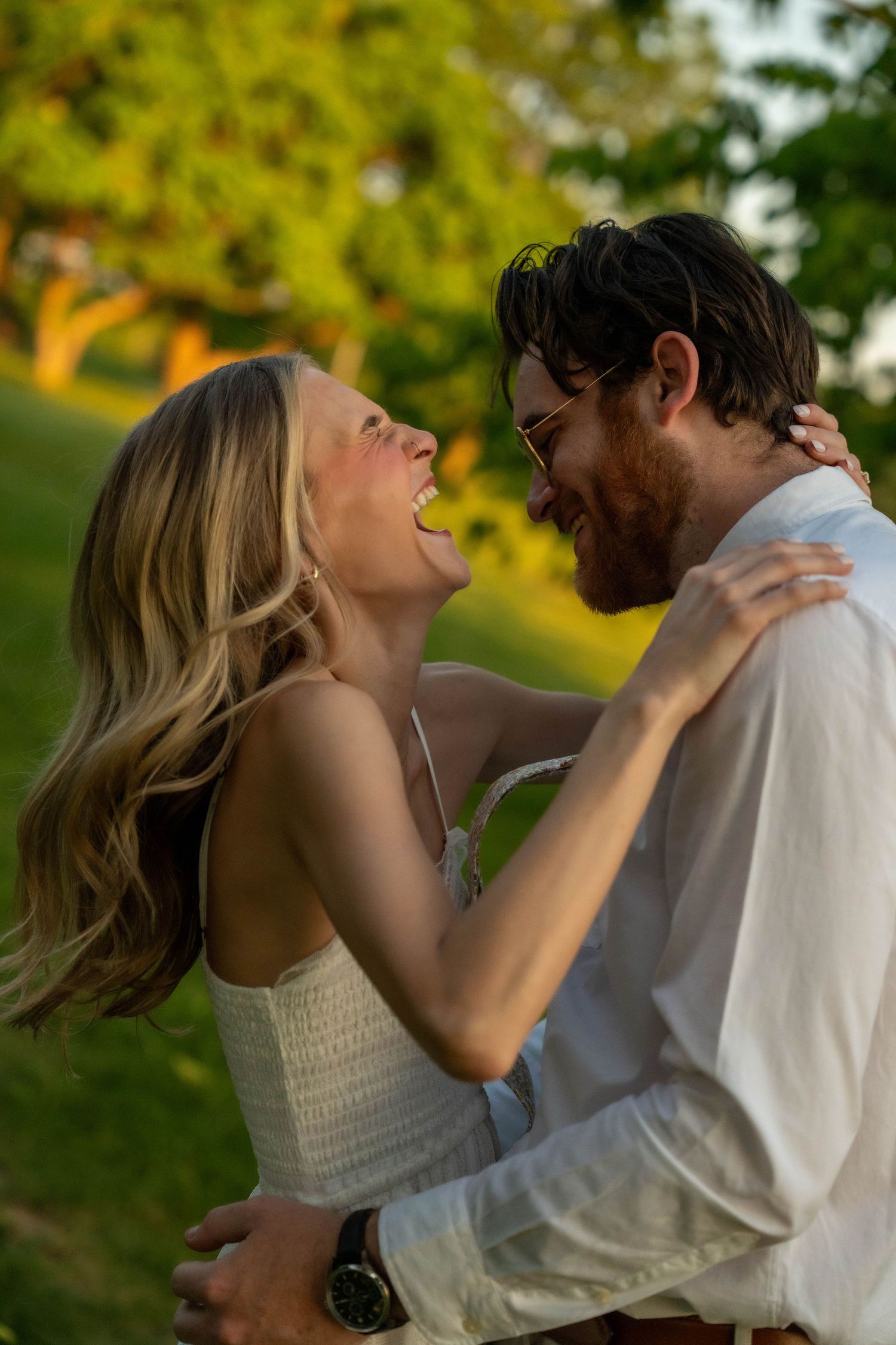 Laughing couple embracing outdoors at sunset, woman in white dress with arms around mans shoulders.