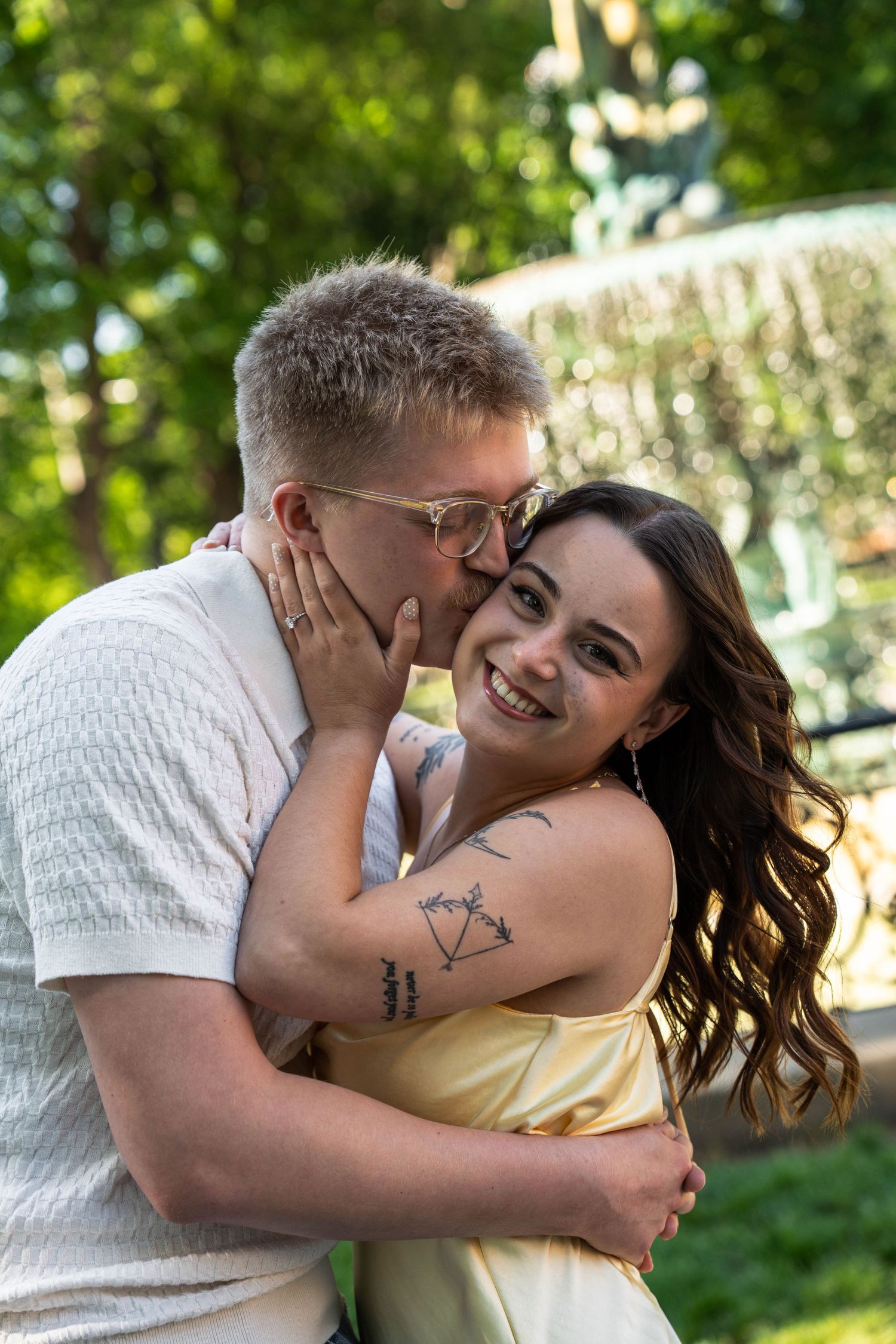 Couple embracing outdoors, man kissing woman's cheek as she smiles, fountain visible in background.
