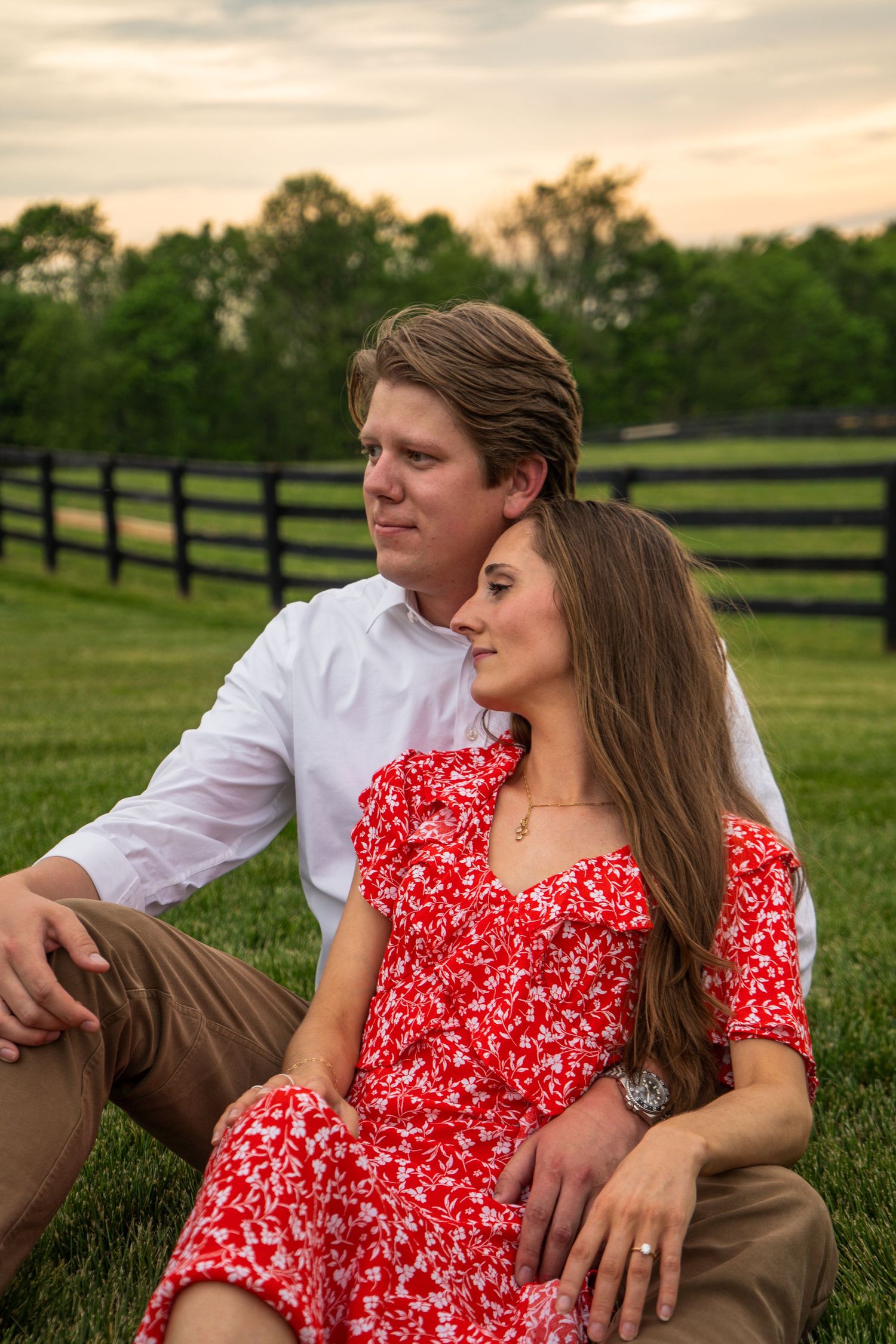 Engaged couple sitting on grass at sunset near black fence, woman in red dress leaning on man.