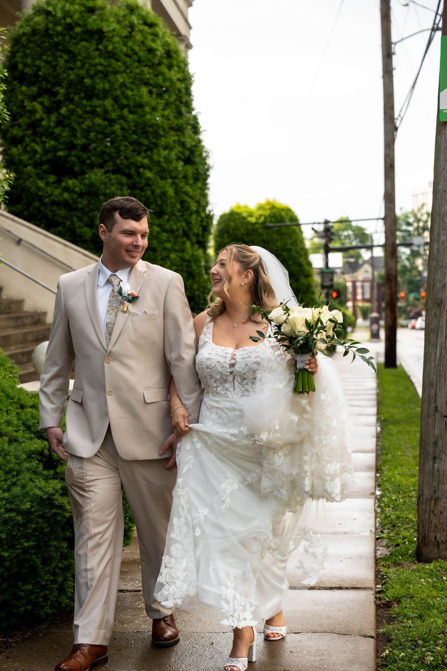 Bride and groom walking hand in hand on a sidewalk, laughing together on their wedding day.