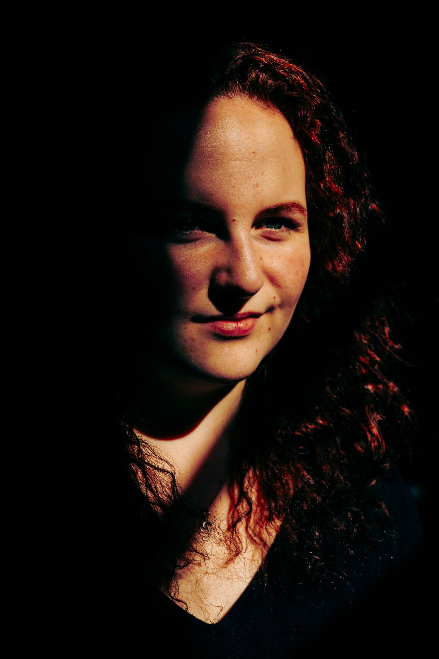 Curly redhead woman smiling softly with dramatic side lighting highlighting her features against dark background.