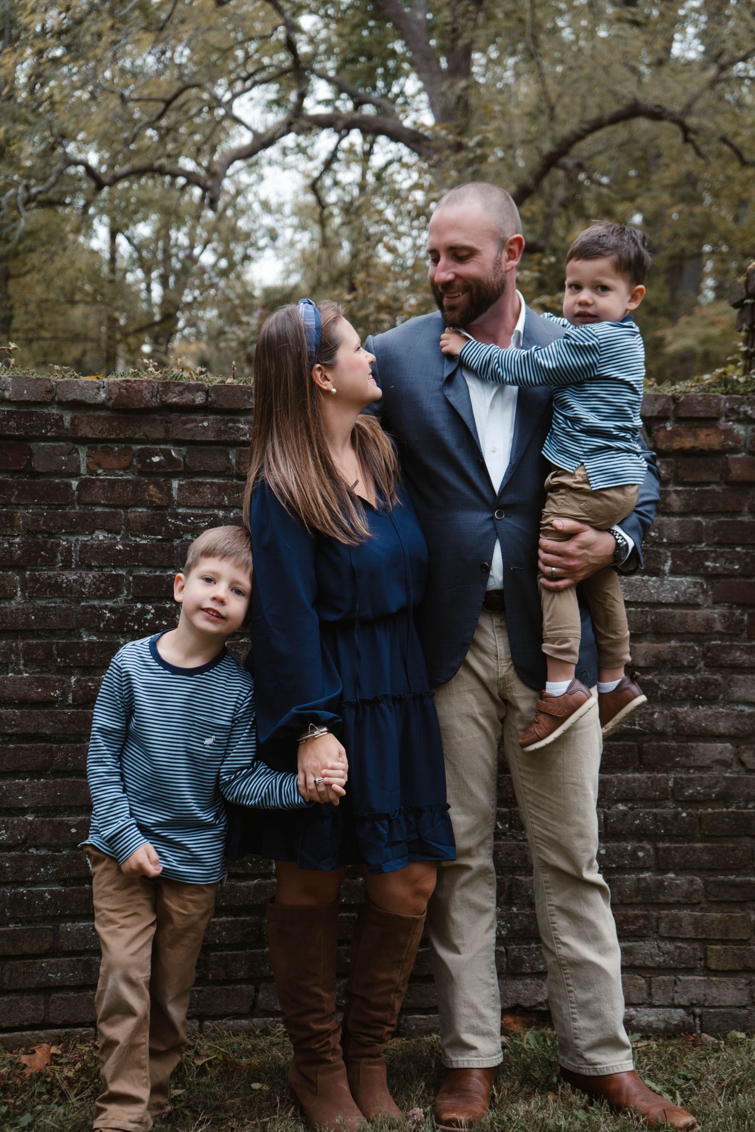 Loving family of four stands together near rustic brick wall outdoors, father holding toddler while mother and son smile.