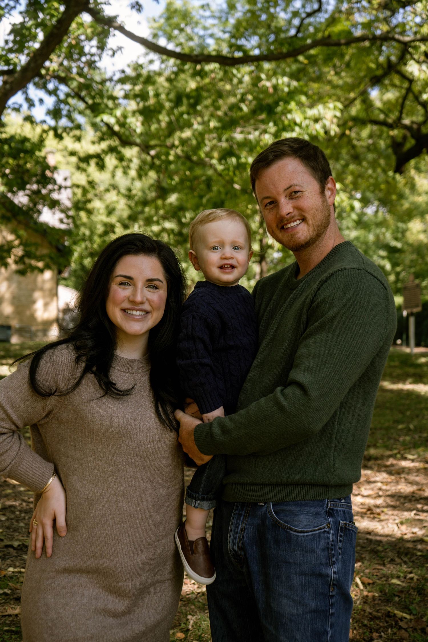 Happy family of three smiling outdoors, dad holding blonde toddler in navy sweater under green trees.
