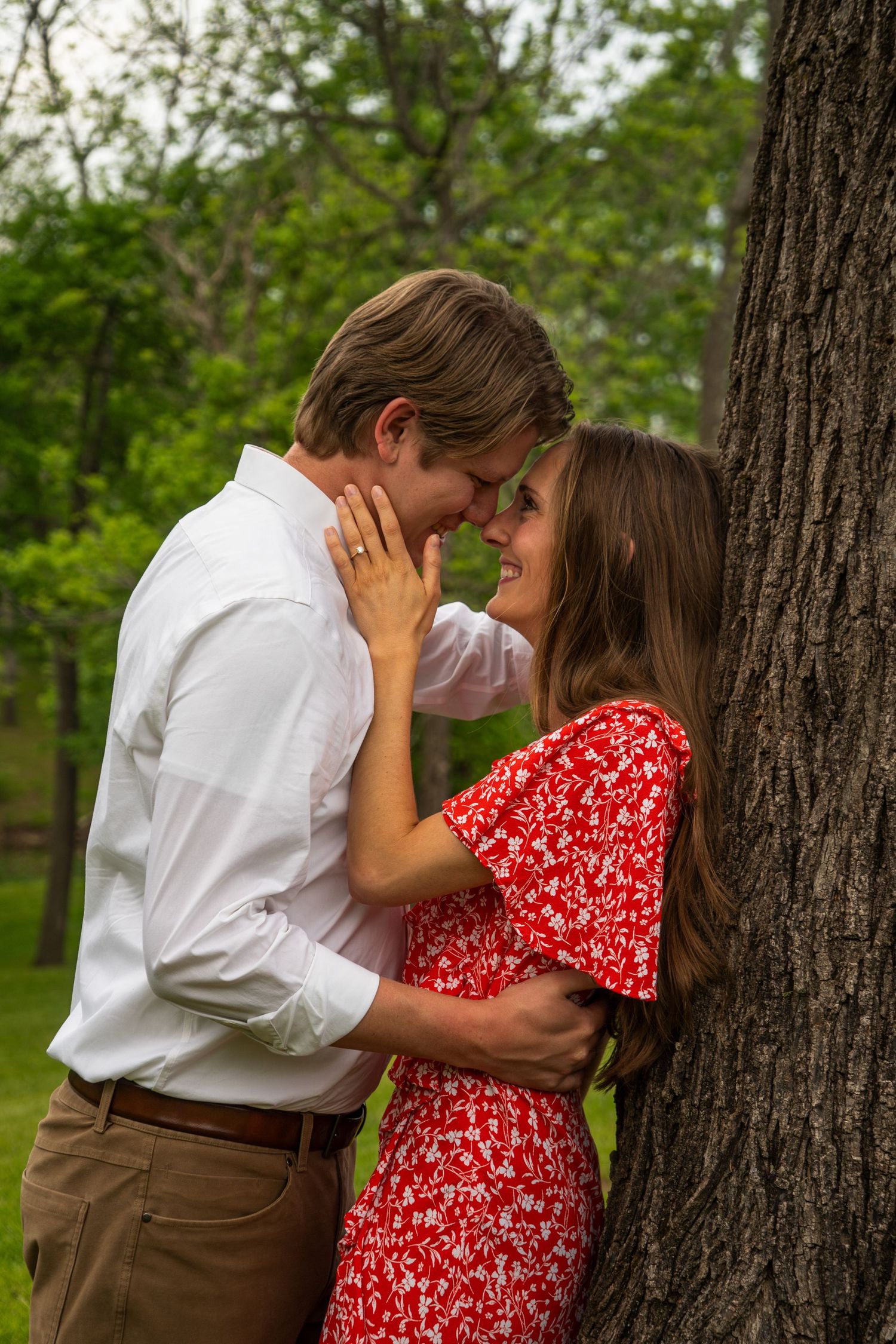 Couple sharing a romantic moment leaning against a tree in a lush green park, woman wearing red floral dress.