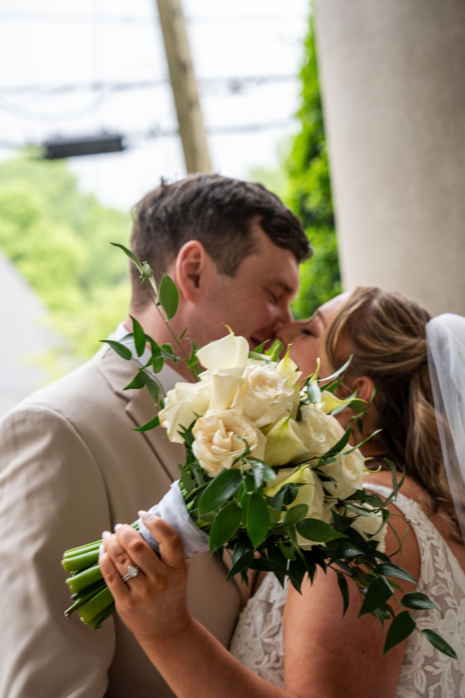 Bride and groom share a kiss behind a bouquet of white roses and greenery at their wedding.