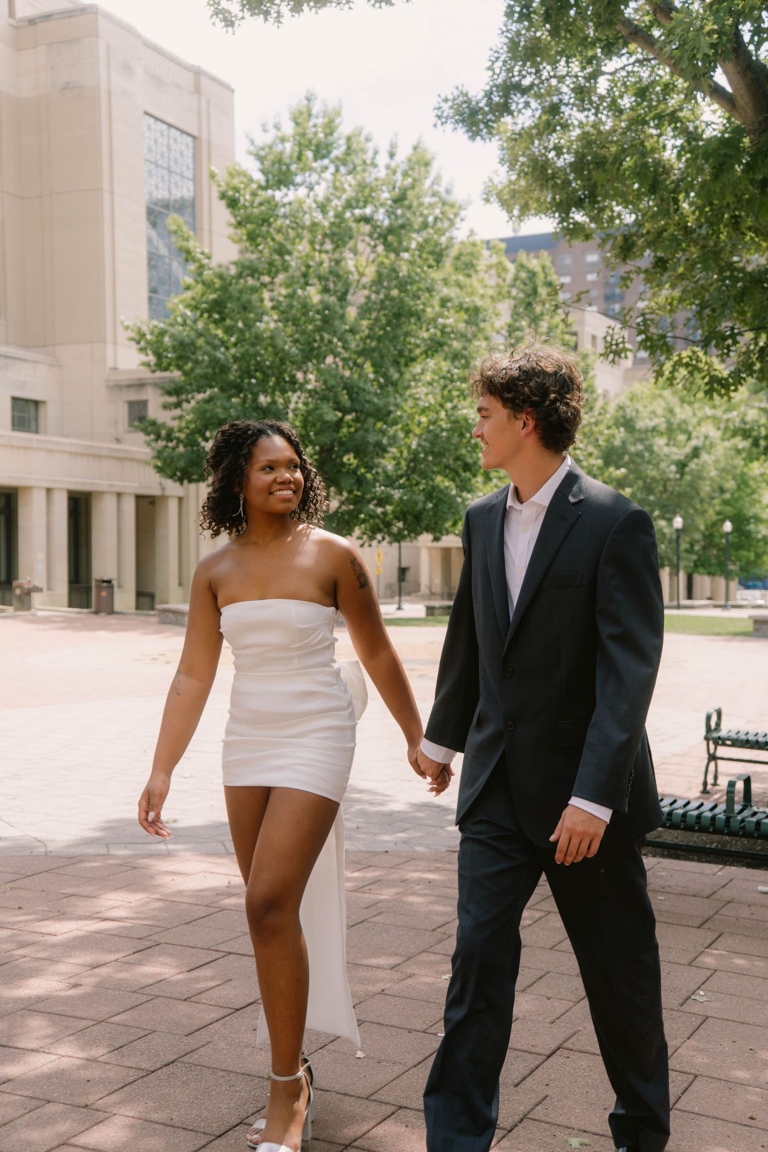 Smiling interracial newlywed couple holding hands walking through sunny outdoor plaza with lush green trees.