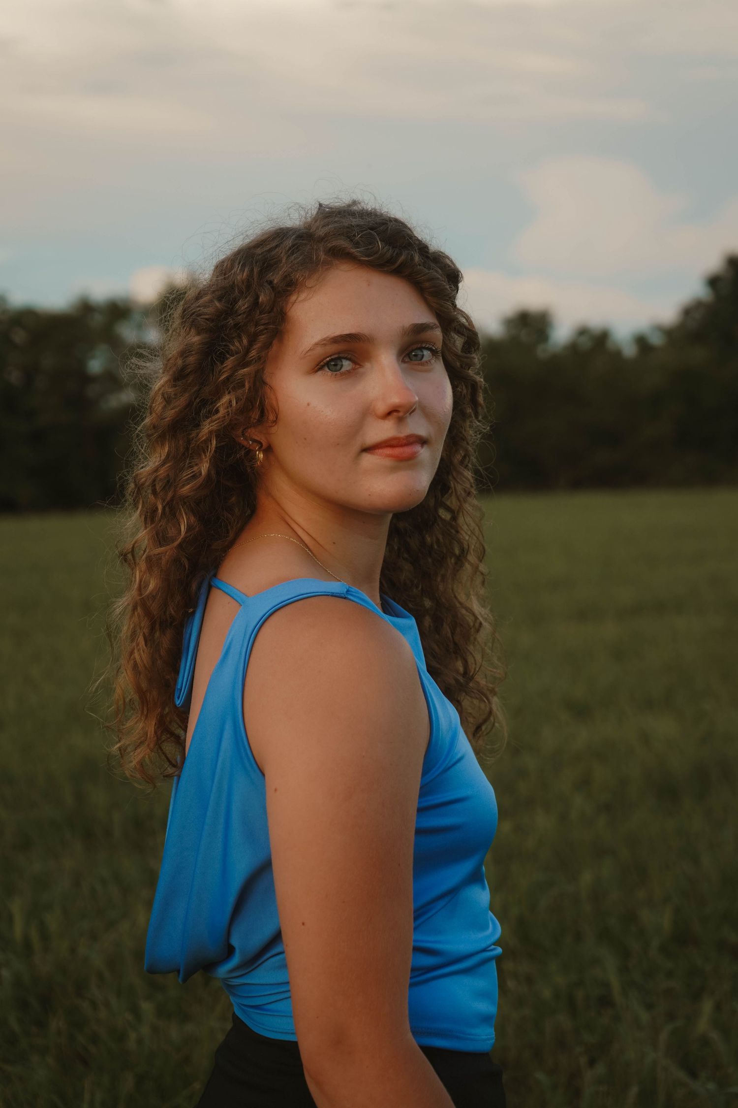 Young curly-haired woman in a blue sleeveless top posing outdoors in a green field at sunset.