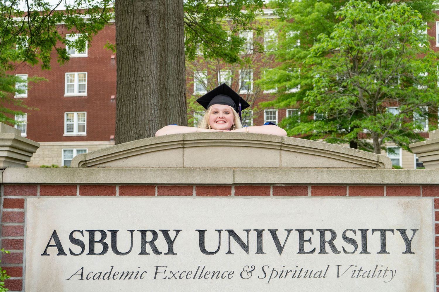 Smiling graduate peeking over Asbury University sign on campus.