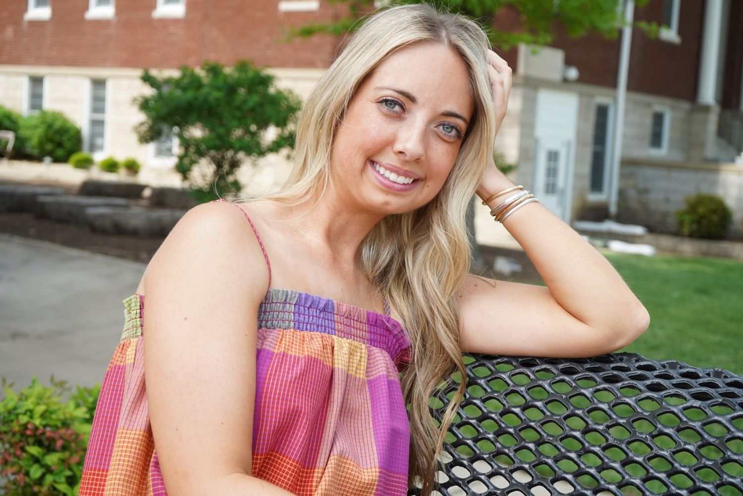 Cheerful blonde woman in pink plaid off-shoulder top sitting outdoors at a bench.