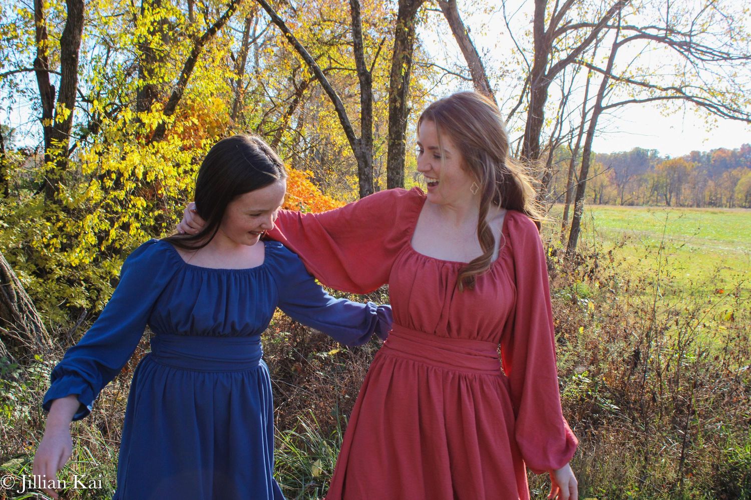Two young women in blue and coral dresses walking in autumn field.