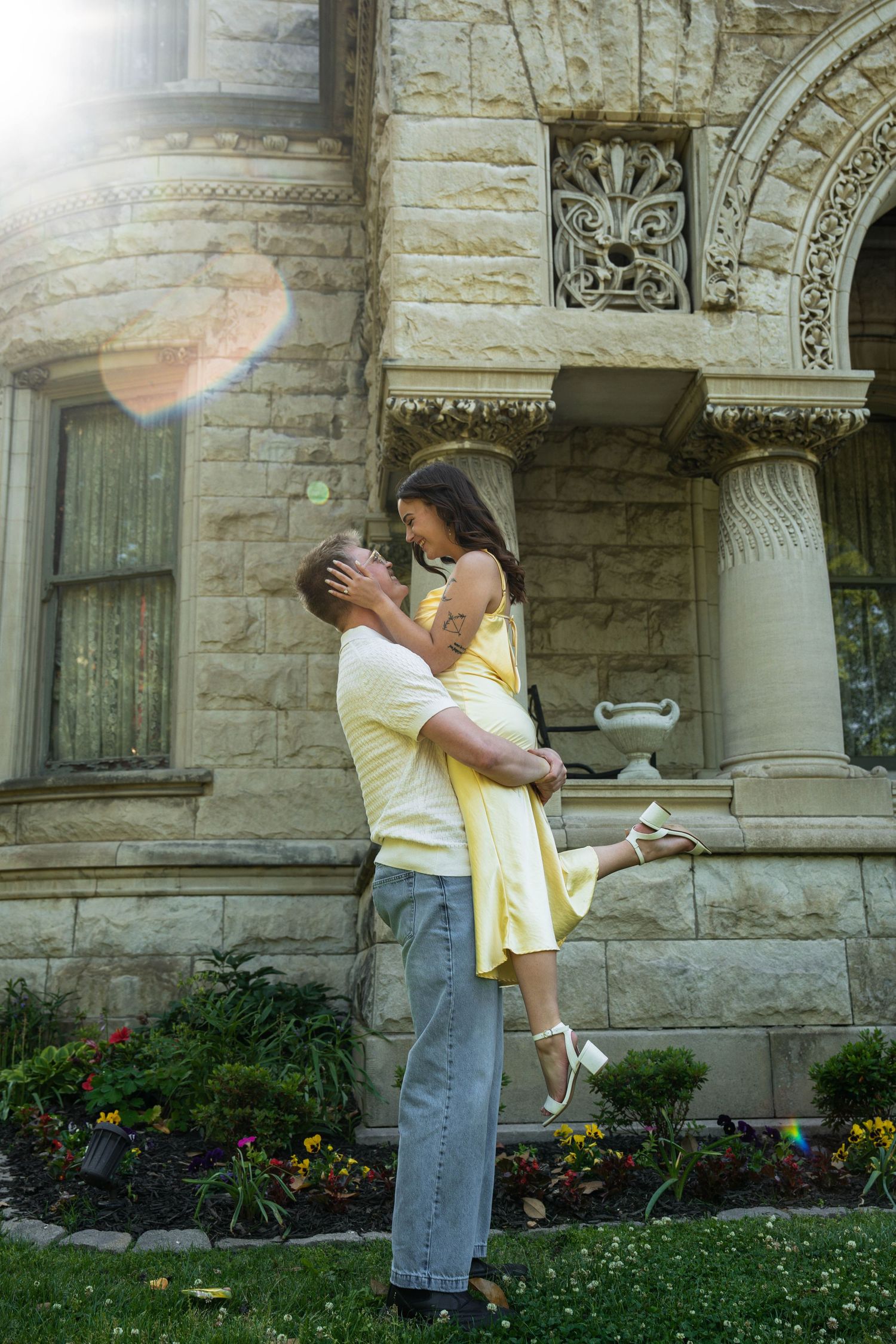 Man lifting woman in yellow dress outside an ornate stone building in sunlight.