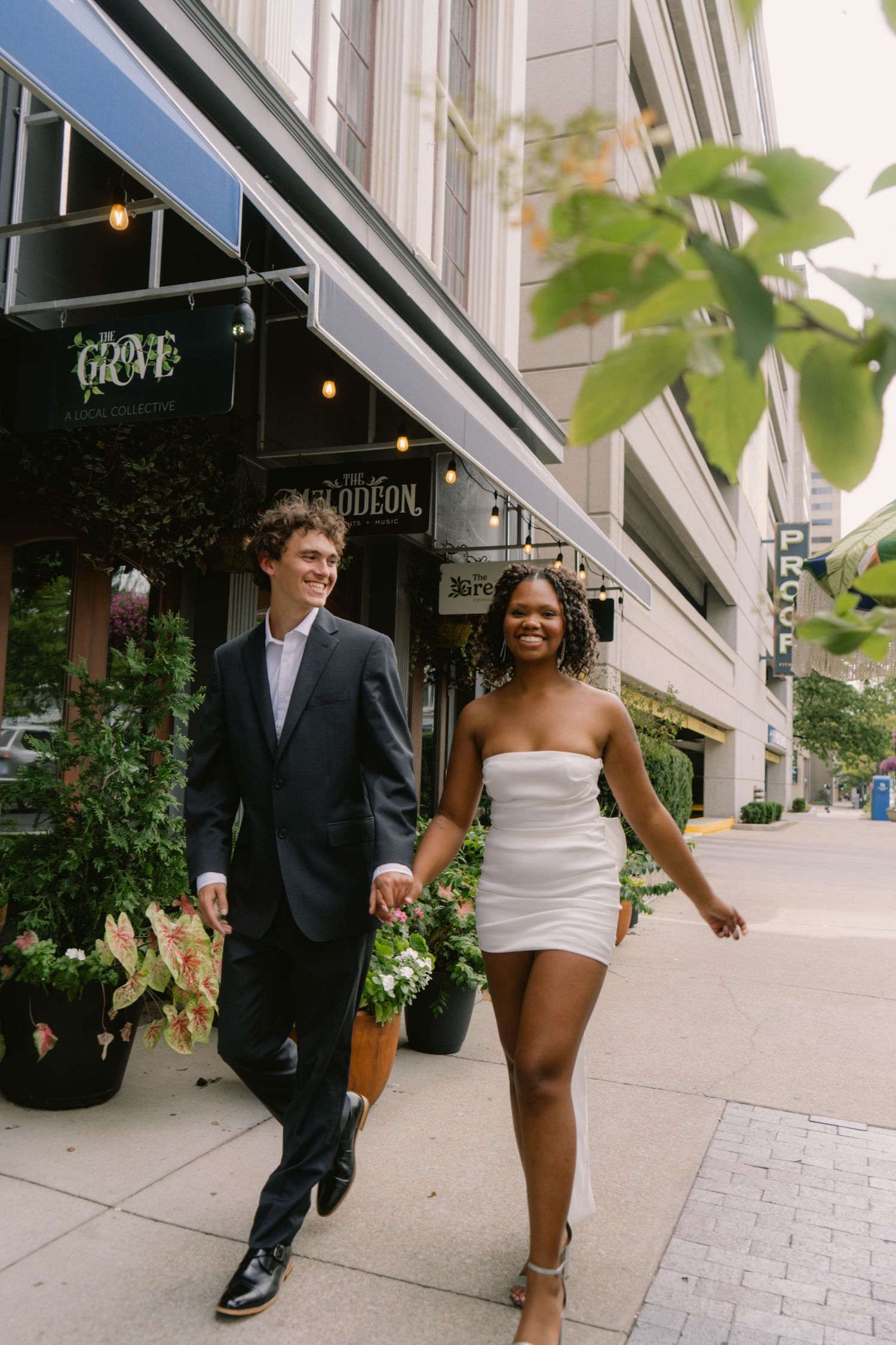Couple in formal wear holding hands and smiling while walking on a city sidewalk past boutique storefronts.