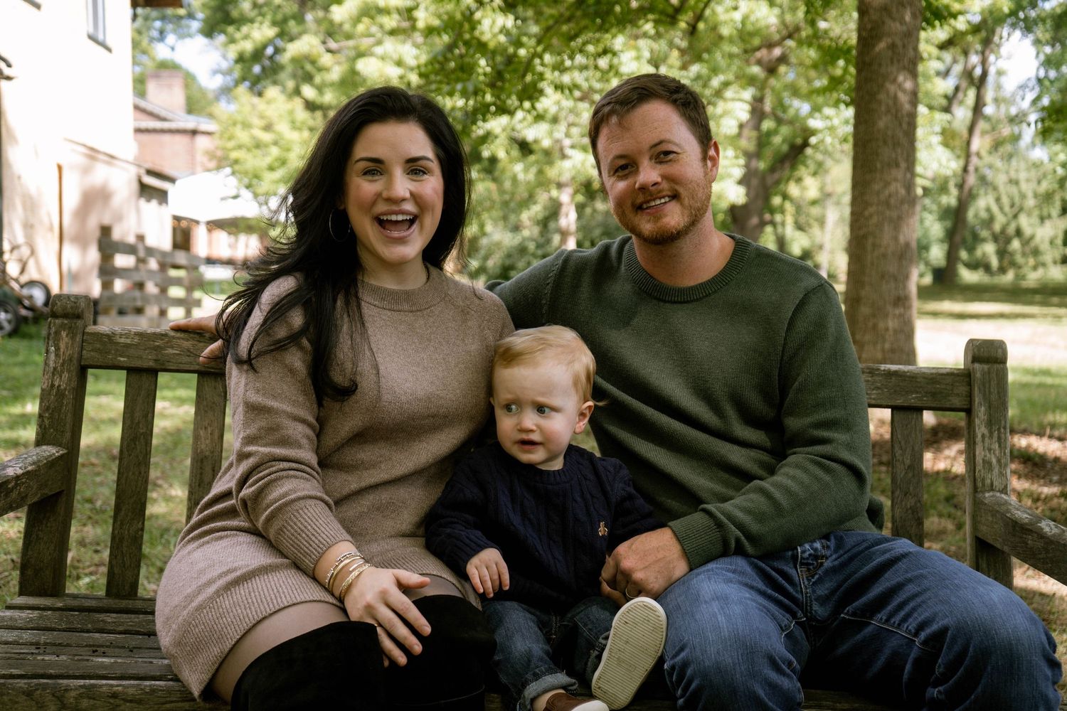Happy young family of three sitting on a wooden bench outdoors, laughing together in a park.