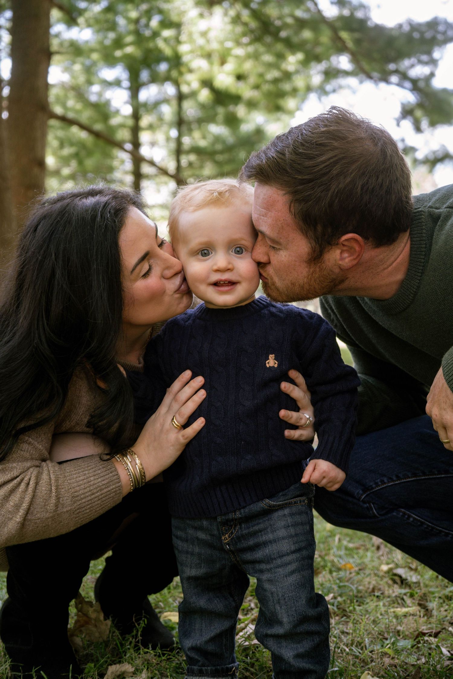 Parents simultaneously kissing their happy blonde toddler on both cheeks while crouching in a park.