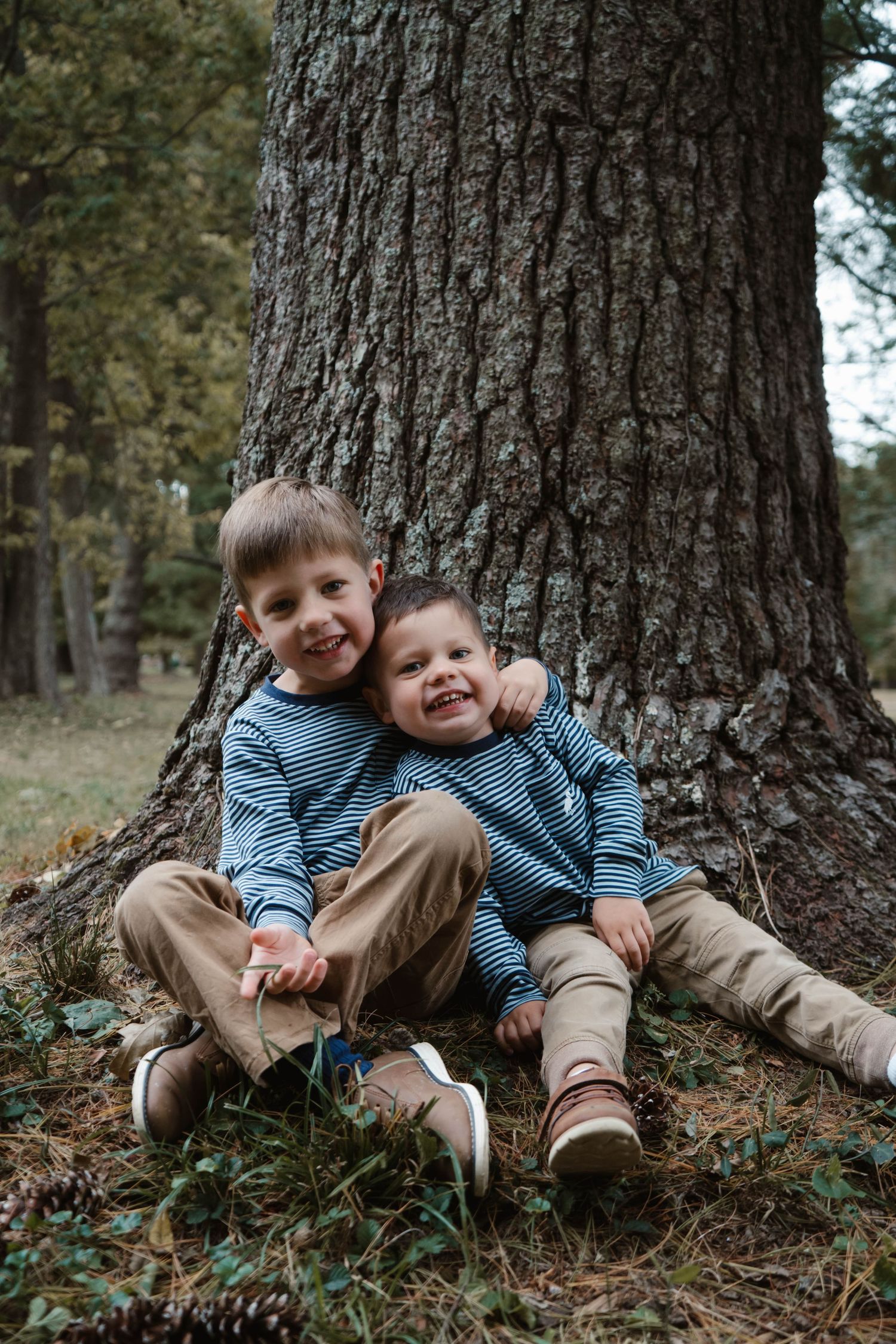 Two young brothers in matching striped shirts smile while sitting together against a large tree trunk in a forest.