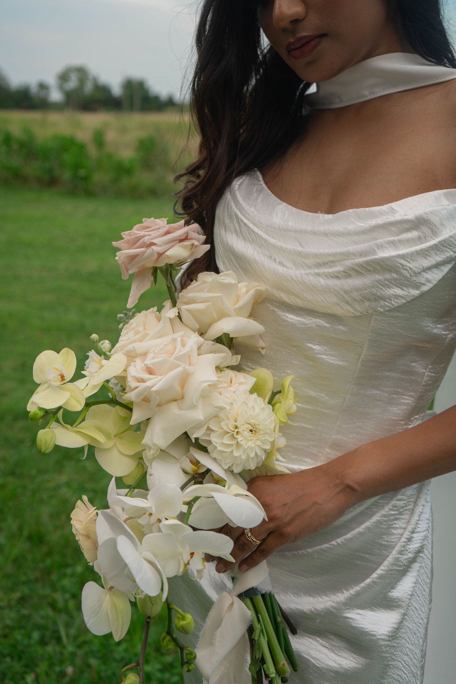 Bride in white off-shoulder gown holds elegant bouquet of blush roses, white orchids, and dahlias outdoors.