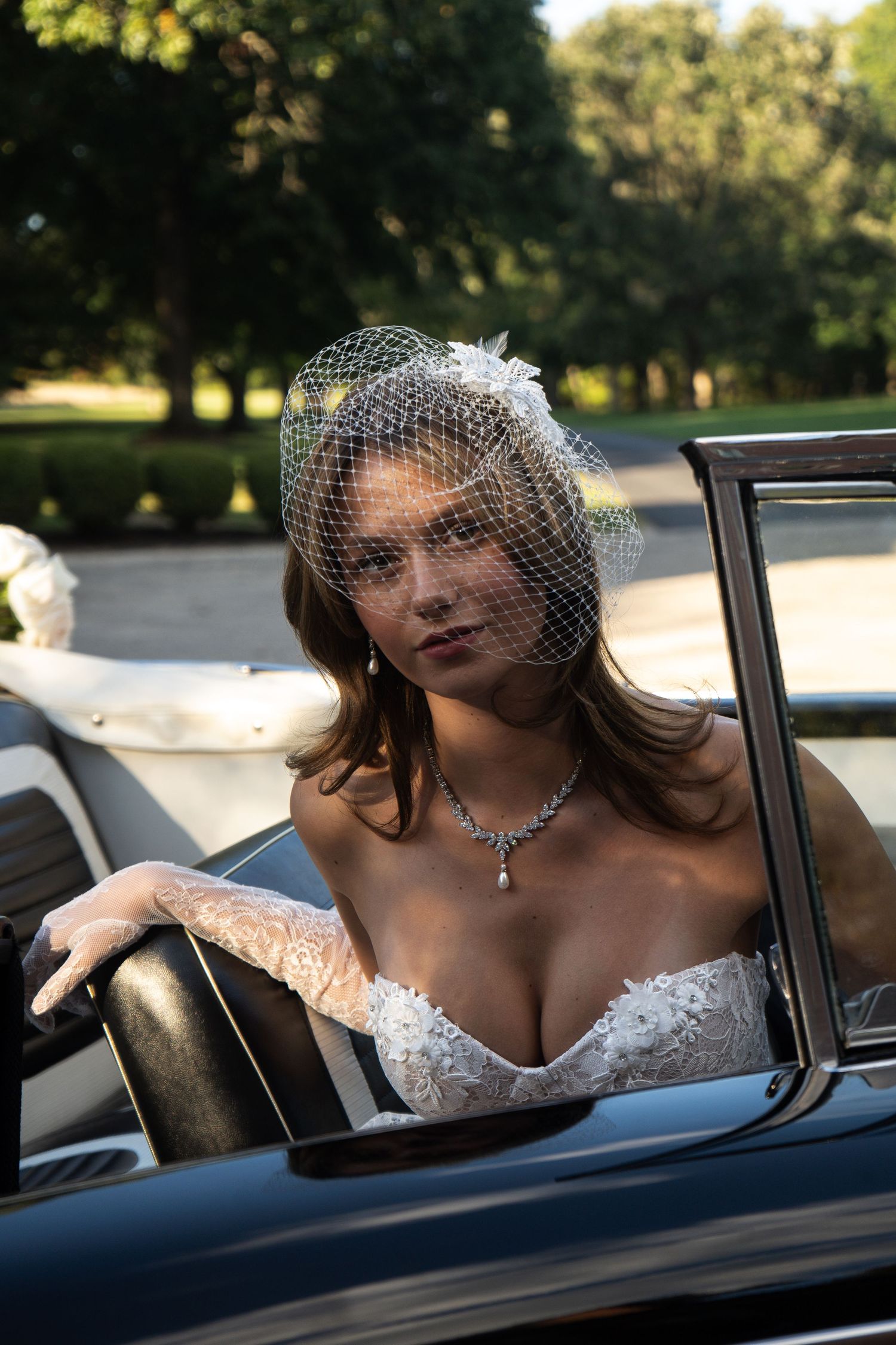 Elegant bride in lace gown and birdcage veil poses in a vintage convertible car outdoors.