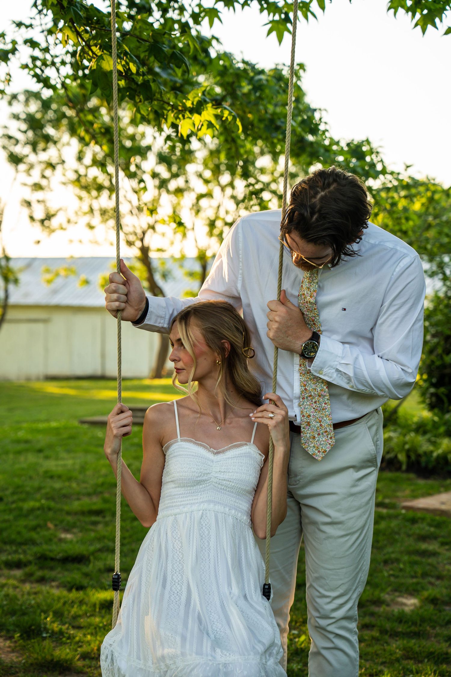 Bride sits on a garden swing in white dress while groom in white shirt leans over her lovingly.