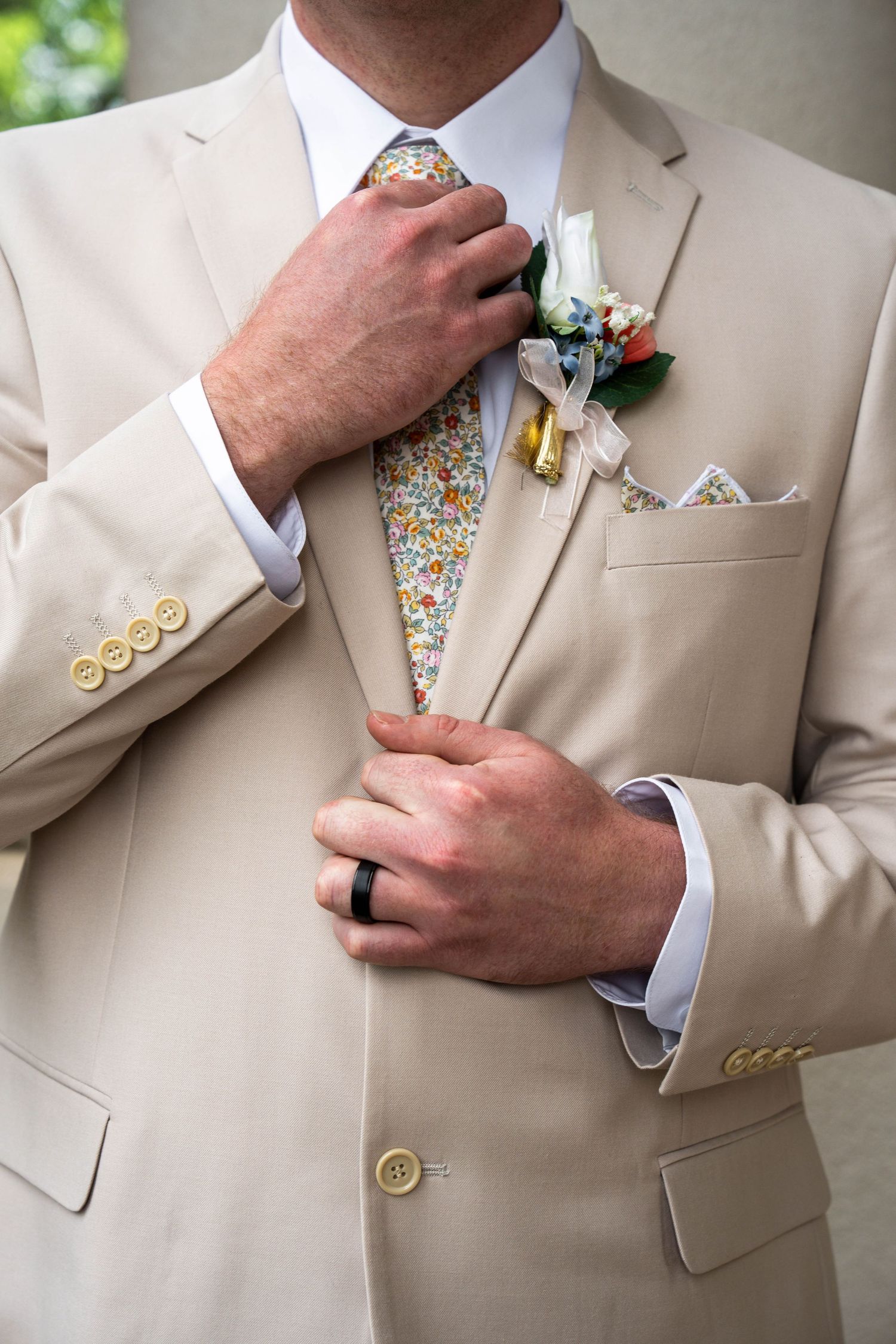 Groom in tan suit adjusts his floral tie, boutonniere and pocket square visible on lapel.
