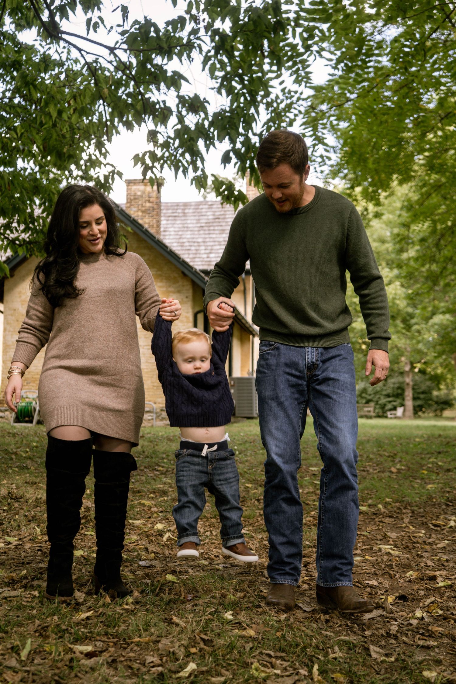 Parents joyfully lift their toddler by the hands while walking in a lush green park setting.
