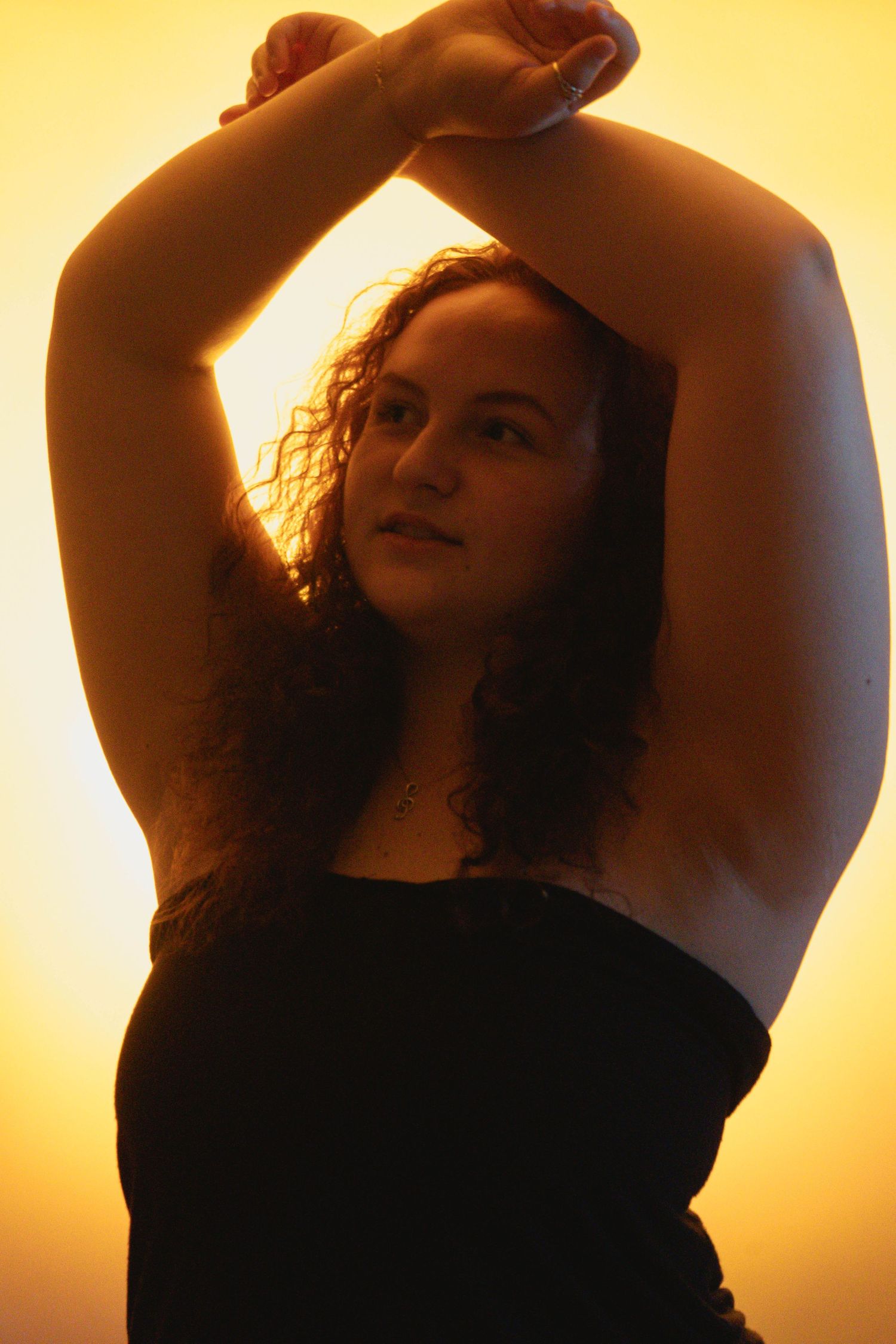 Woman in black top posing with arms raised overhead against a warm golden yellow backlit background.