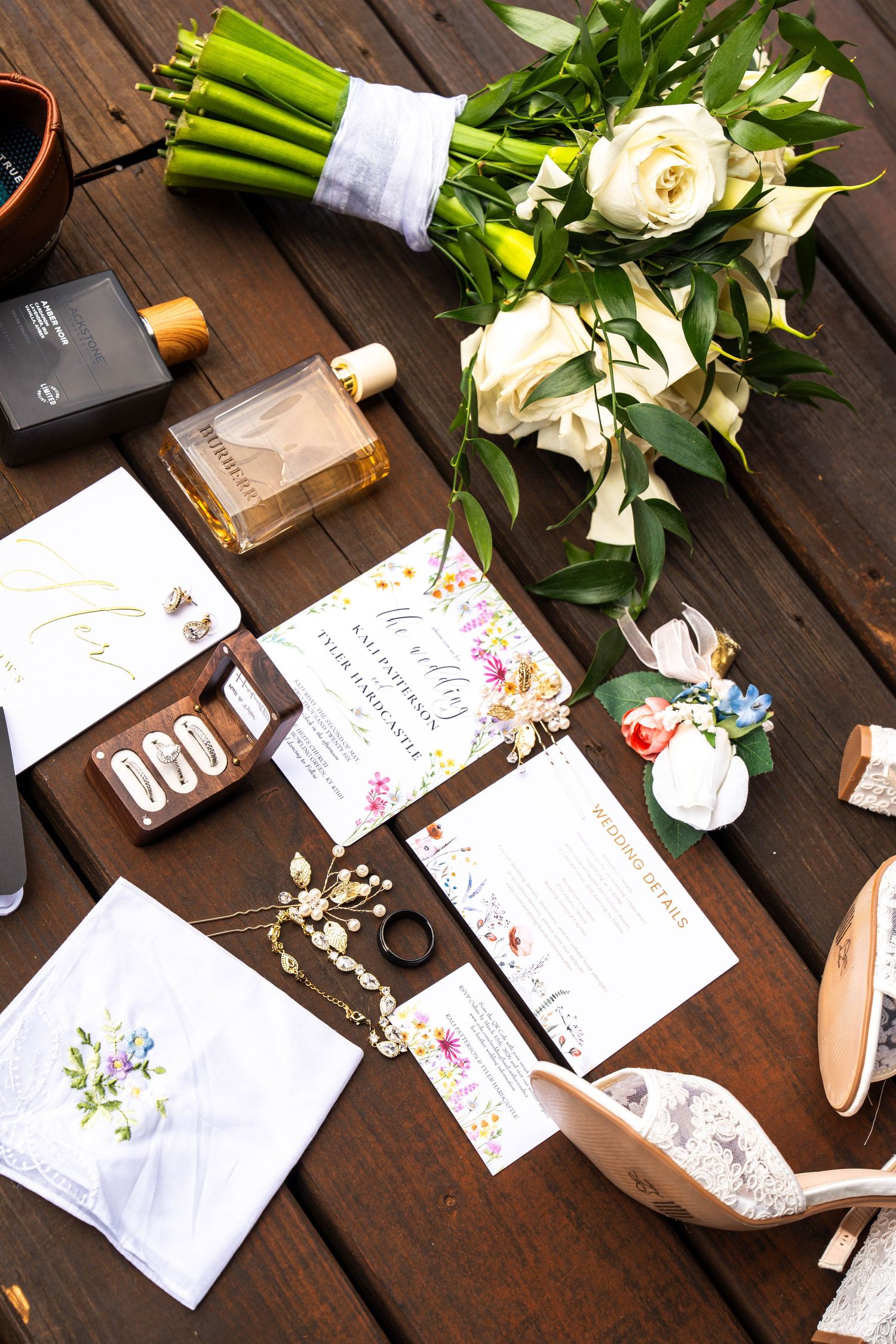Flat lay of wedding stationery, white roses bouquet, perfume, shoes, and accessories on wood table.