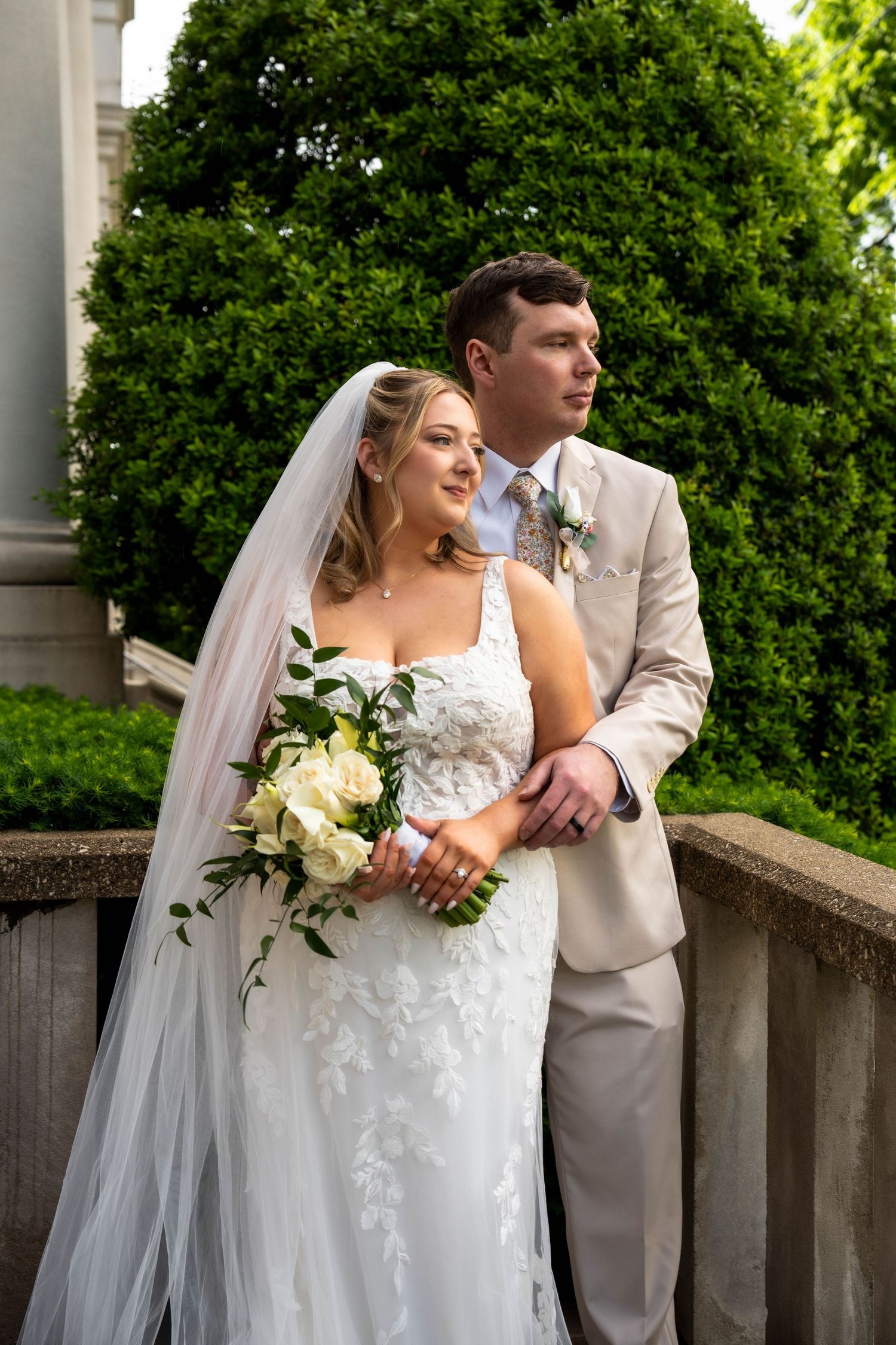 Bride in lace gown with veil and groom in tan suit pose together on stone balcony with greenery.